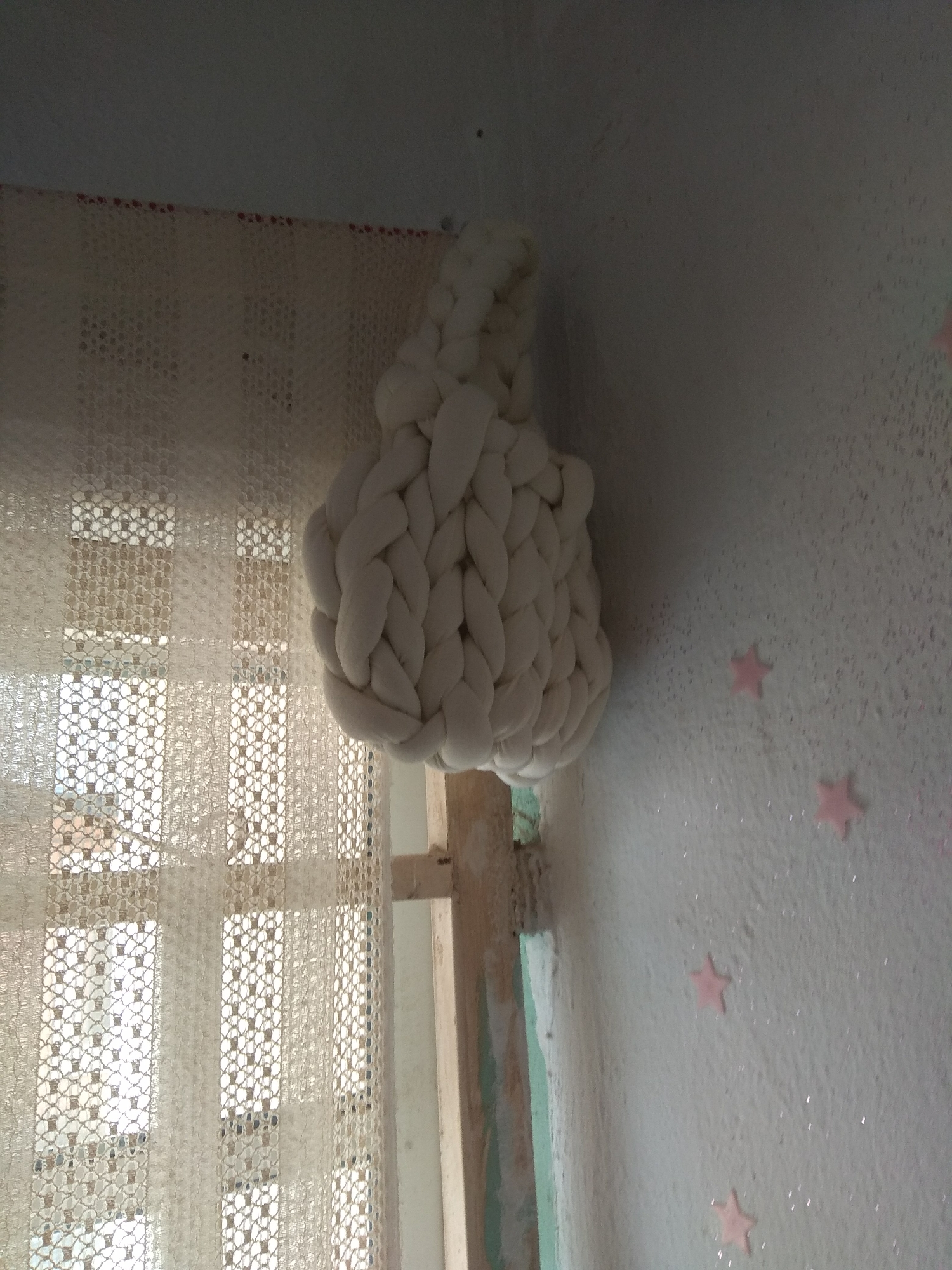 This is a medium-angle, eye-level shot, taken indoors, likely in a residential setting, depicting a wall and a window. The wall on the right is textured and painted white, with small pink star-shaped decorations scattered across its surface, some of which appear to have a glitter finish. A thick, cream-colored, braided object, possibly made of yarn or fabric, hangs from the ceiling on the left side of the frame, obscuring a portion of a white lace curtain that covers a window. The window is visible through the translucent curtain, revealing multiple panes. The environment appears to be a room in Jalingo, Nigeria.