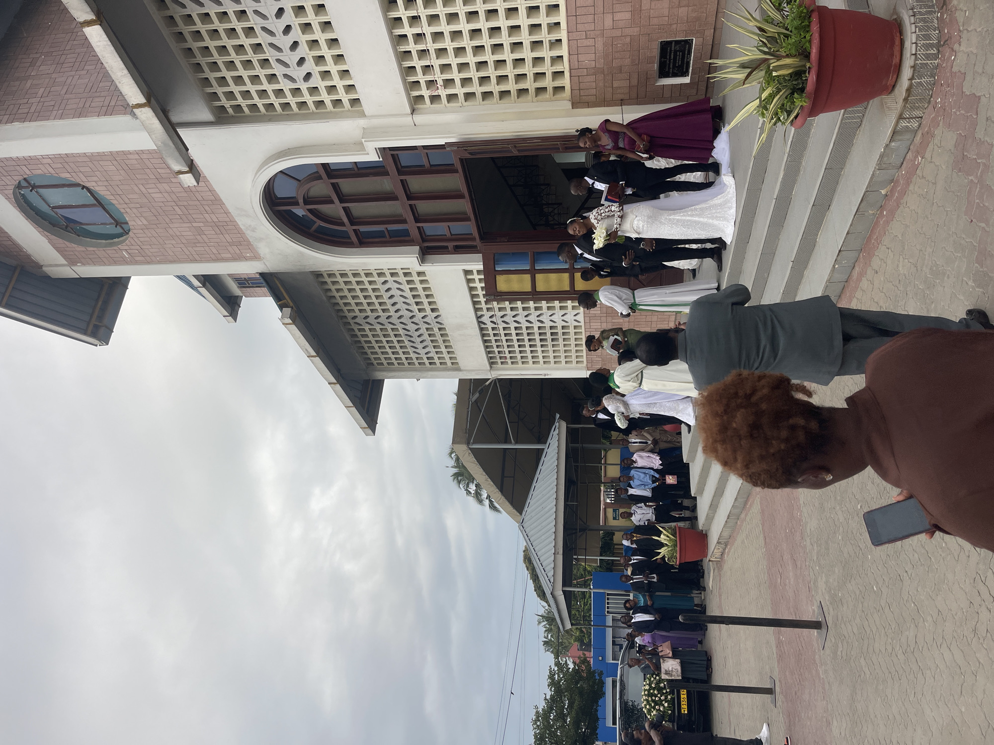 In the city of Dar es Salaam, Tanzania, a wedding ceremony is taking place outside a church. The bride, wearing a white lace gown, stands with her groom, who is dressed in a black tuxedo. They are surrounded by wedding guests, some of whom are dressed in formal attire. The church has a distinctive architectural style, with a pink tiled facade and a large arched doorway. A black car decorated with flowers is parked in the foreground, possibly belonging to the wedding party. A woman in a brown shirt is holding a phone, appearing to take a photo or video of the event. The sky is overcast, suggesting a cloudy day. The overall scene depicts a joyful and celebratory occasion, with a sense of community gathered to witness the union of the couple.