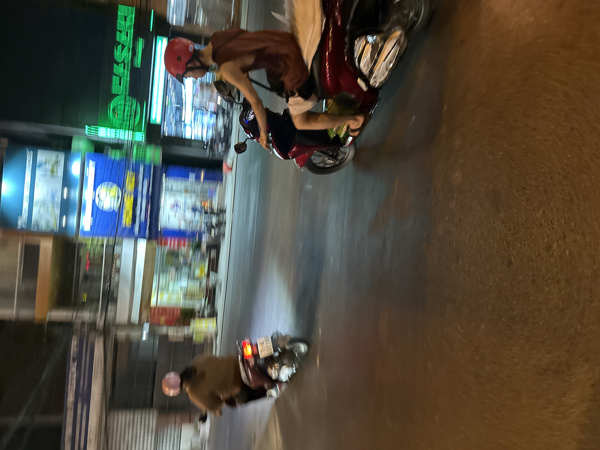 The image depicts a street scene in Bac Nha Trang Ward, Vietnam at night. Two people are riding motorbikes. The first person wears a red helmet, brown shirt, and light-colored shorts, riding a red motorbike. The second person wears a brown jacket, riding a dark-colored motorbike. Streetlights and illuminated signs are visible in the background.