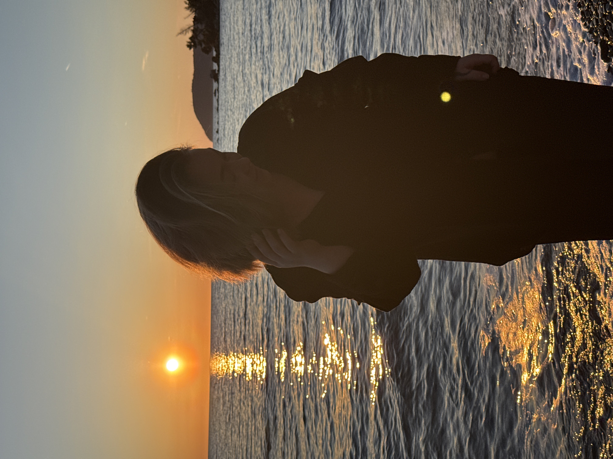 A long shot of a person standing on the shore of a beach, with a sunset in the background. The person is silhouetted against the bright light of the sun. They are wearing a black jacket. They have their hand up to their face as if they are in deep thought. The water is calm, with the sun reflecting off the waves. The sky is a clear blue, with a few clouds in the distance. The location is the city of Bar, Montenegro.