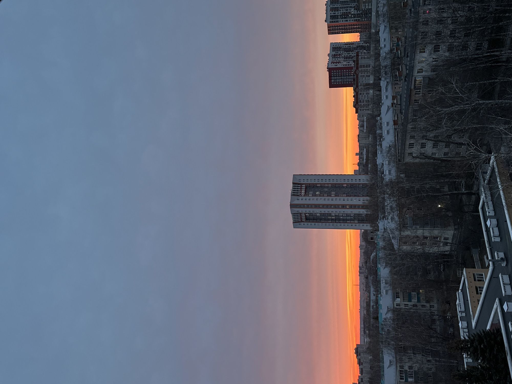 The image captures an urban scene at sunset, possibly in Kazan, Russia, given the architecture. The sky transitions from a dark blue-gray to orange near the horizon. Several high-rise buildings and apartment blocks are visible, some with red accents. Bare trees and rooftops are present, with some evidence of snow or frost on the structures. The photograph shows a city skyline at dusk, emphasizing the contrast between natural and man-made structures.