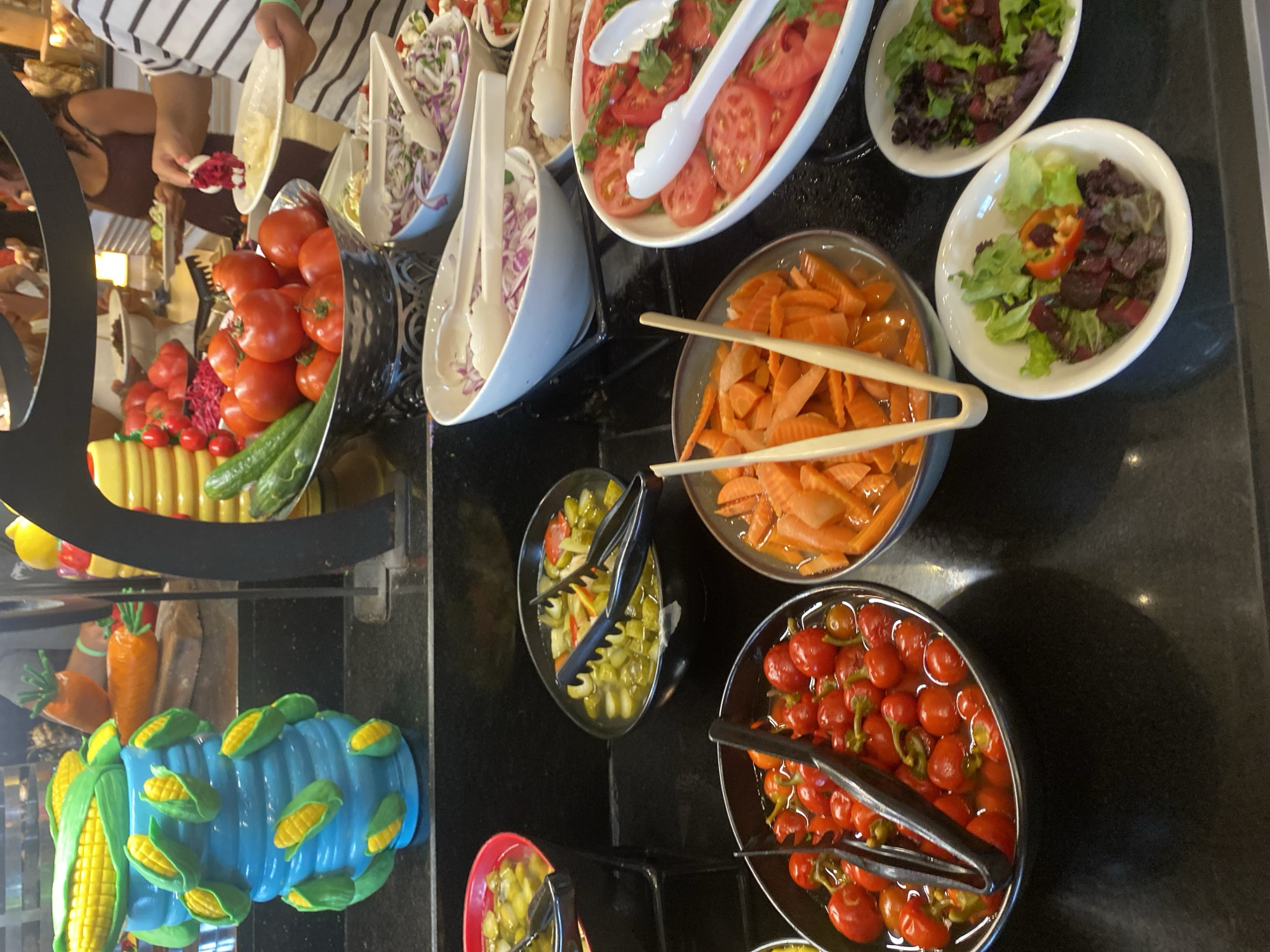 An indoor buffet in Aksu, Turkey, features a variety of fresh foods. In the foreground, a dark bowl overflows with pickled cherry peppers in brine, with black tongs resting inside. To its right, a bowl of sliced carrots in liquid contains light-colored tongs. A white oval dish to the right holds sliced tomatoes with a sprig of parsley, and next to it, a smaller white oval dish contains a salad with lettuce and diced beets, garnished with a slice of red pepper. A larger white oval dish further back contains a salad with lettuce and diced beets, with a slice of red pepper visible. Another white oval dish holds sliced red onions and some greens. A silver bowl in the middle background is filled with whole tomatoes and cucumbers. To the left, a red circular dish contains sliced pickles in brine, with black tongs. Behind these, a blue decorative vase shaped like a corn cob holder is adorned with several smaller corn cob decorations. Further back, blurred figures of people are visible, some holding plates and interacting with the buffet. The environment appears to be a well-lit dining area with dark countertops.