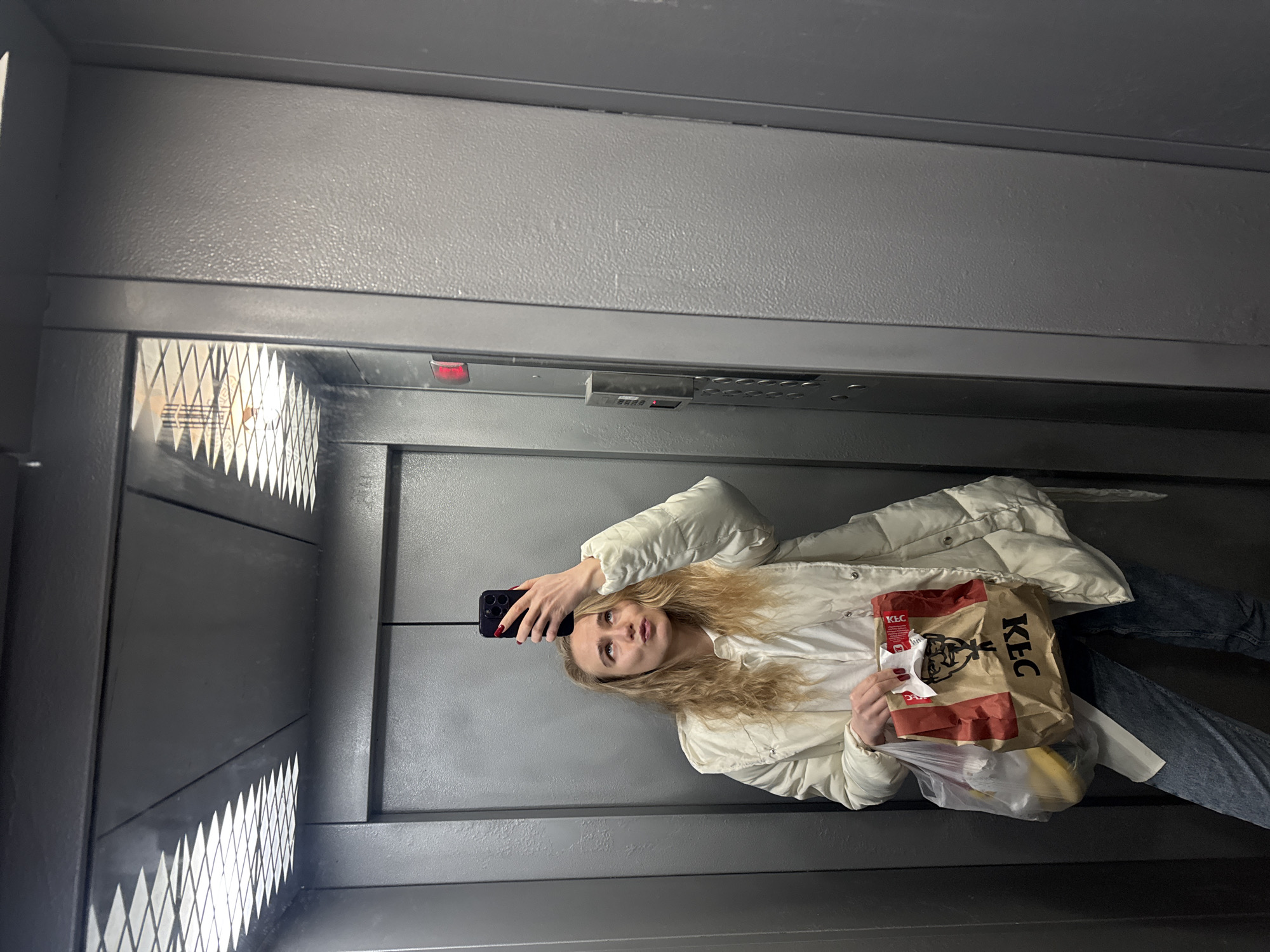 A woman takes a selfie in an elevator, holding a KFC bag. She wears a white puffer jacket, a white shirt, and blue jeans. The elevator interior is gray with horizontal lines and a light panel. Location: Unknown, Unknown.