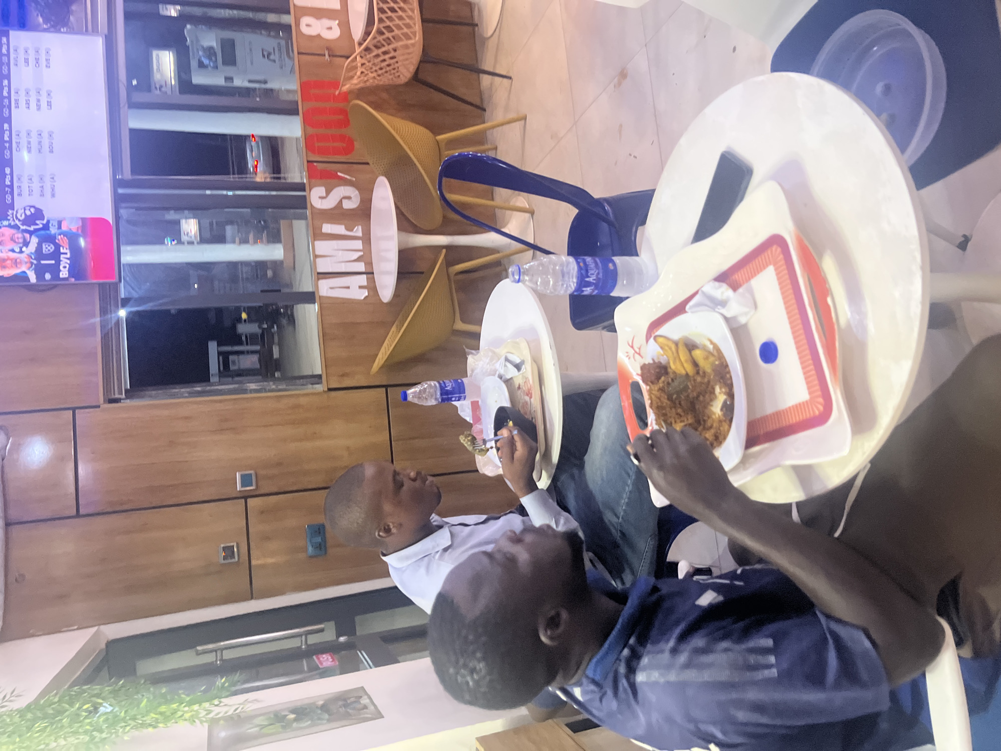 Two dark-skinned men are eating at a restaurant. The image is rotated counterclockwise, which affects the perspective and framing. One man, wearing a white shirt, is seated to the left, facing another man who is wearing a blue striped shirt. Both are eating from plates on white tables. The tables are set with trays that contain food (likely fried yam and stew) and bottled water. The environment suggests a commercial establishment, indicated by the display of Premier League sports on a TV screen in the background, furniture, and decorations. The walls have wooden paneling, and the furniture includes yellow and blue chairs. The sign indicates "Am-St. and Com &," which is likely the name of the restaurant and confirms the location as Abuja, Nigeria.