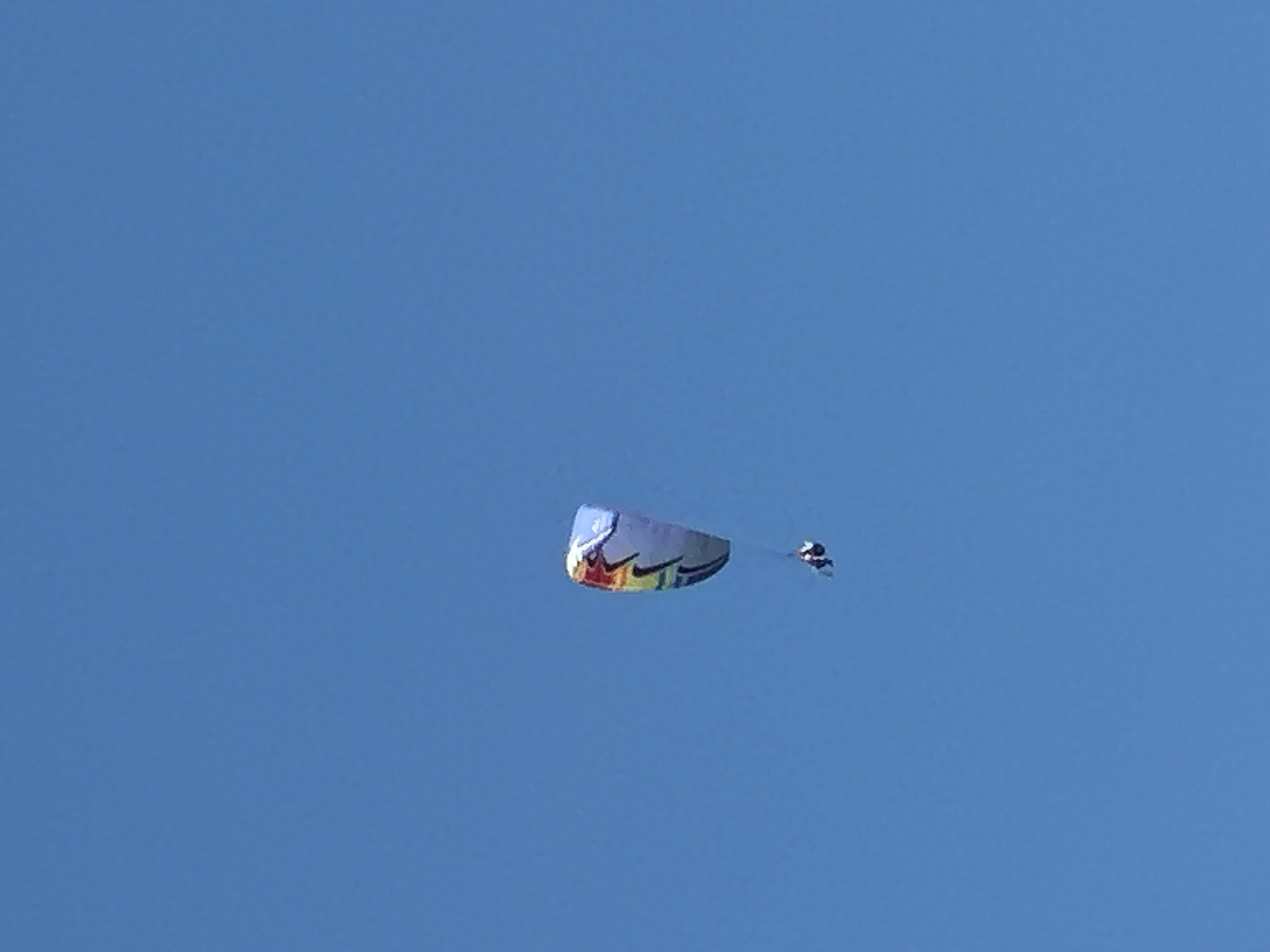 A paraglider is captured mid-flight against a clear blue sky. The paraglider itself is colorful, featuring stripes of red, yellow, green, and blue against a white background. A person is visible suspended beneath the paraglider, appearing to be in control of the descent. The image does not provide any specific information about the location in Boreti, Montenegro.
