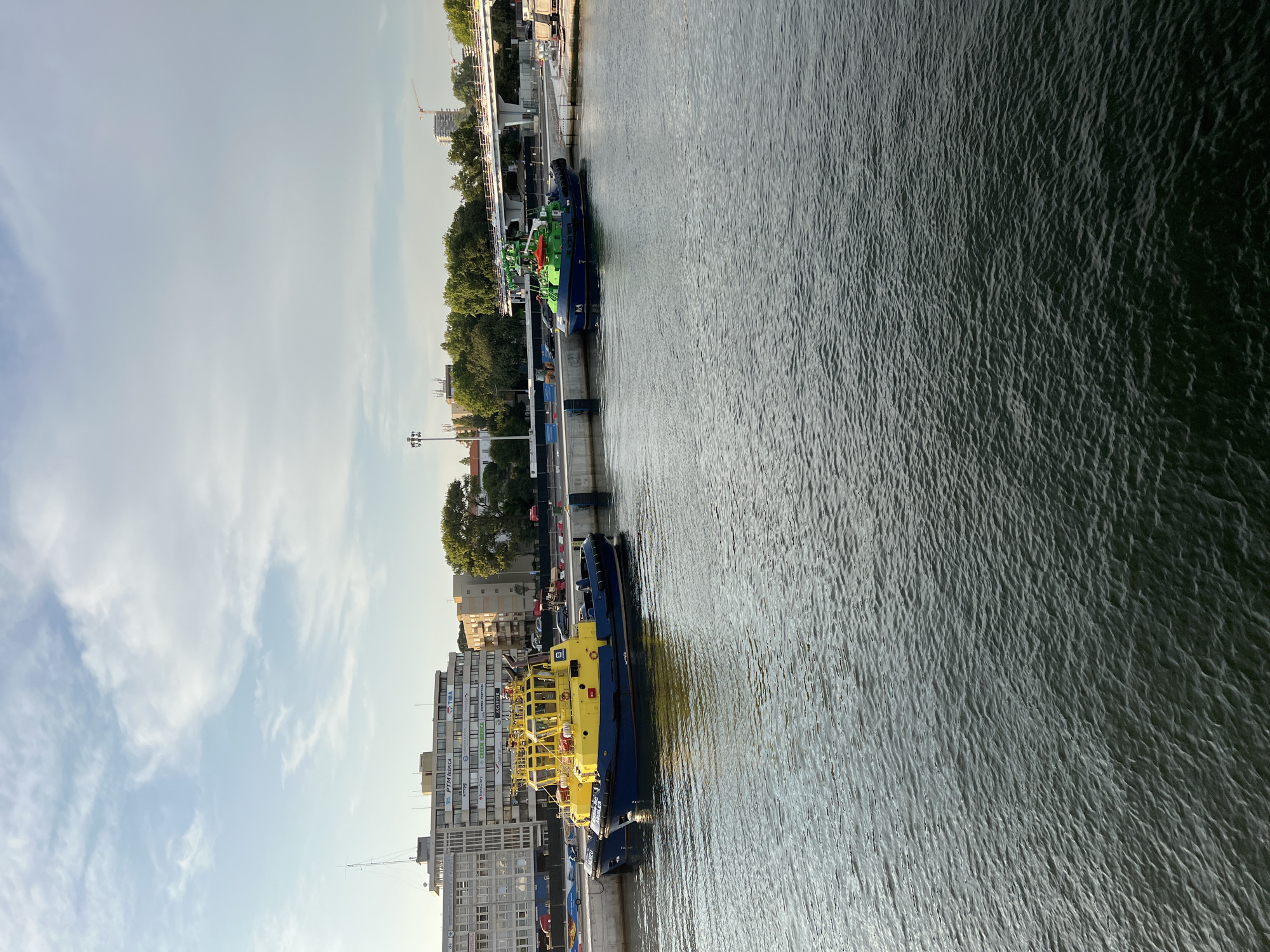 The image shows a harbor in Matosinhos, Portugal, with several tugboats docked at a pier. The tugboats are yellow and blue, and green and blue, respectively. In the background, there is a long, multi-story building with several signs, indicating commercial activity. Trees and a bridge are also visible in the background, suggesting an urban or semi-urban environment. The water in the foreground is dark and appears calm, with small ripples on the surface. The sky is partly cloudy, with a mix of blue sky and white clouds. The lighting suggests it is either morning or late afternoon. There are no people visible in the image.