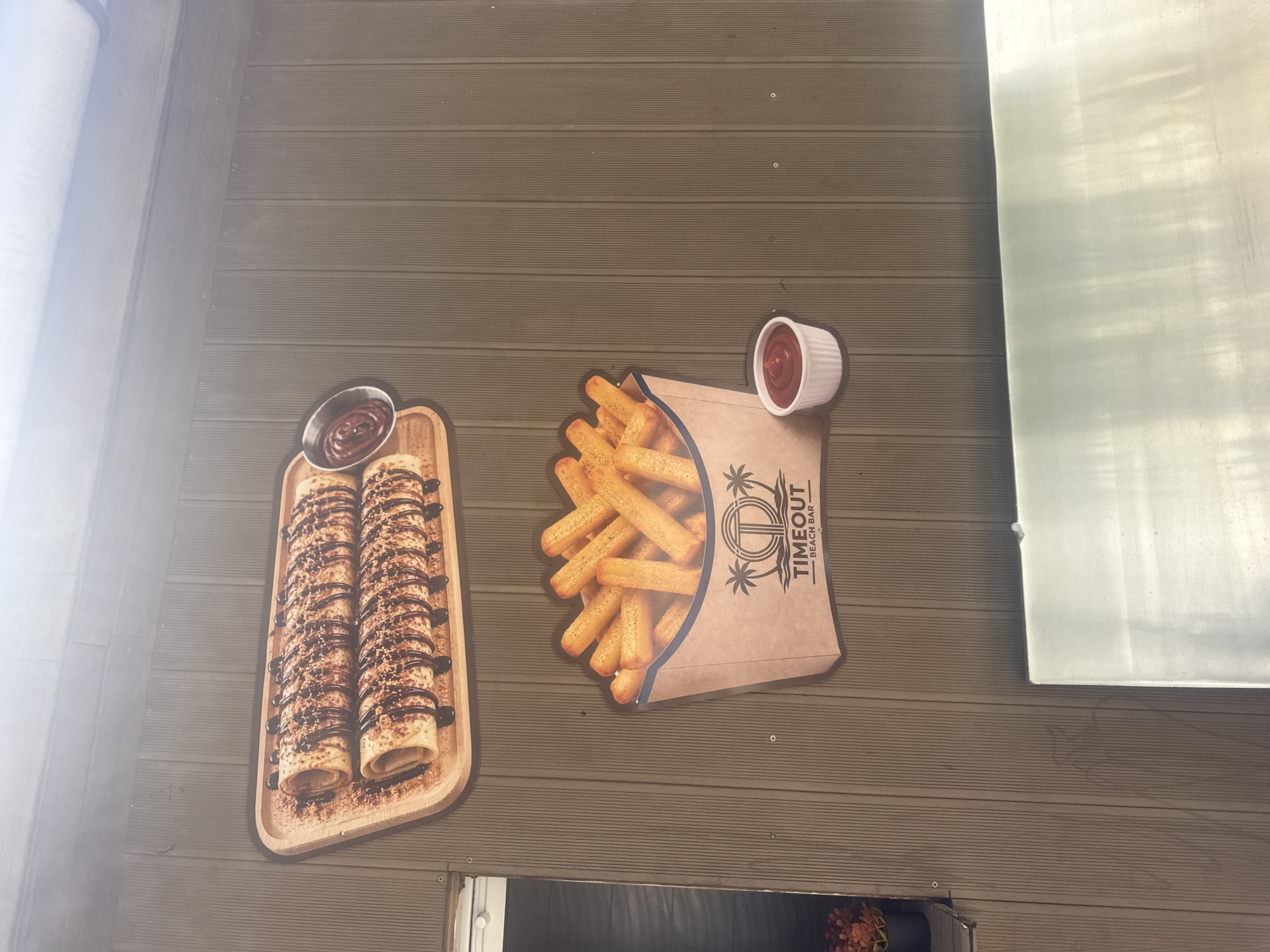 A media file displays two signs affixed to a wall made of horizontal wooden planks. On the left, a sign features a wooden tray with two rolled crepes topped with chocolate syrup and what appears to be crushed nuts or cookie crumbs. A small bowl of chocolate sauce sits above the crepes.
To the right, a sign depicts a stylized paper cone filled with golden-brown french fries. A small white ramekin of ketchup is positioned at the top of the cone. The cone has a logo with palm trees and the text "TIMEOUT BEACH BAR".
The wall appears to be an exterior surface, possibly part of a building or kiosk. The lighting suggests it is daytime, with some light reflecting off a large, possibly metallic or mirrored surface to the right of the signs. The overall scene has a casual, potentially beachside or outdoor dining atmosphere, consistent with the "Beach Bar" text. No people are visible in the image.