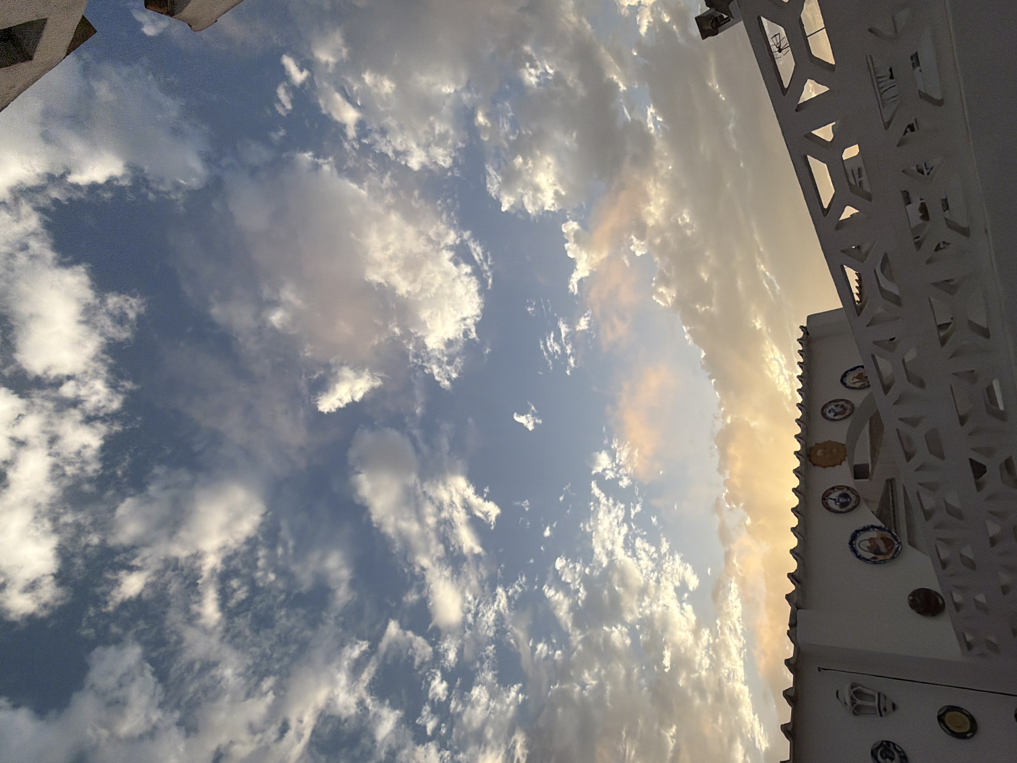 The image depicts a sky filled with a mixture of white and grey clouds, illuminated by a warm, soft light suggesting either sunrise or sunset. Some clouds have an orange or pinkish hue, indicating the presence of the sun low on the horizon, possibly to the right of the frame. In the foreground, the upper section of a white building with decorative elements is visible. These elements include several round ceramic plates, some with painted scenes, affixed to the wall. A white, ornamental fence or railing with a geometric, lattice-like design runs across the lower part of the image. The architectural style of the building and the decorative plates suggest a Mediterranean or Spanish aesthetic. Given the context of the user's request, this scene is likely located in Torrevieja, Spain, a coastal city known for its characteristic white buildings and decorative tile work, often seen along its promenades and residential areas. No people are visible in the image. The environment is characterized by a clear sky with dramatic cloud formations and a hint of architectural detail.