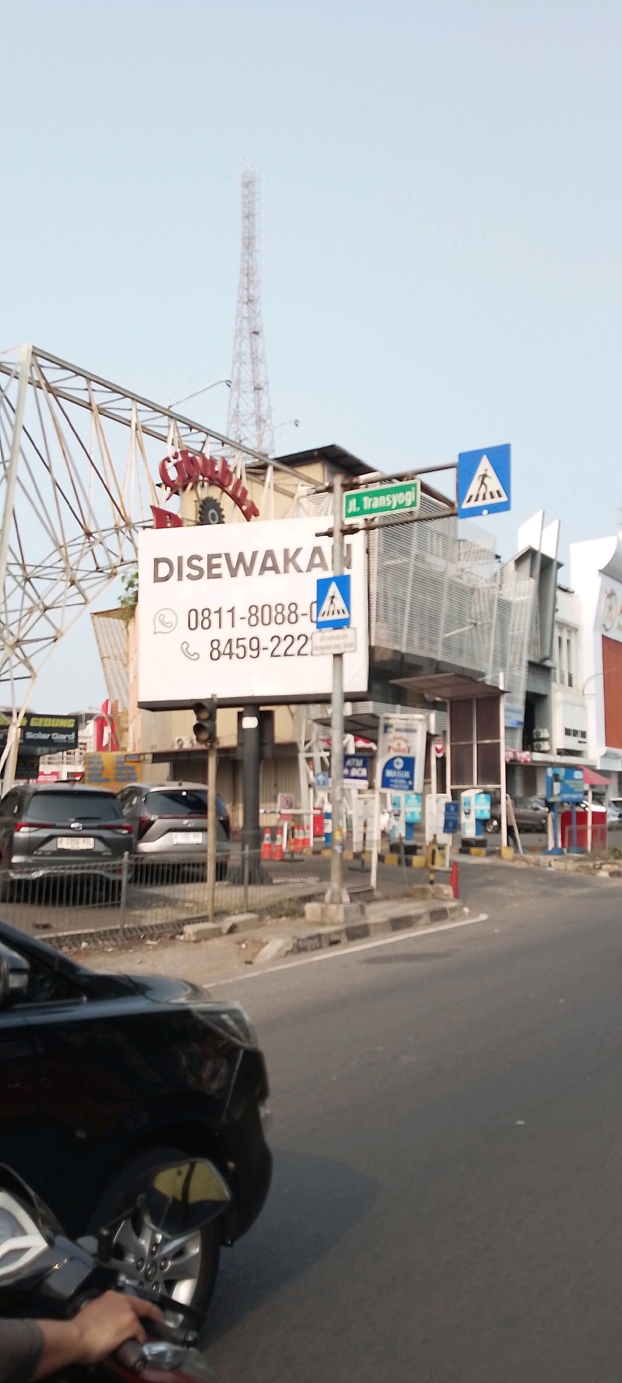 The image captures a street scene in Depok, Indonesia, specifically on Jalan Transyogi. A tall radio tower is visible in the background against a pale sky. In the mid-ground, a large white billboard is prominently displayed, advertising "DISEWAKAN" (for rent) with phone numbers "0811-8088-8" and "8459-222". Adjacent to the billboard are traffic signs, including a pedestrian crossing sign. A street sign indicates "Jl. Transyogi". Several cars are parked behind a fence, and traffic cones are visible further down the road. A black car and a portion of a motorcycle are in the foreground, suggesting the image was taken from a moving vehicle. The environment appears to be an urban commercial area.