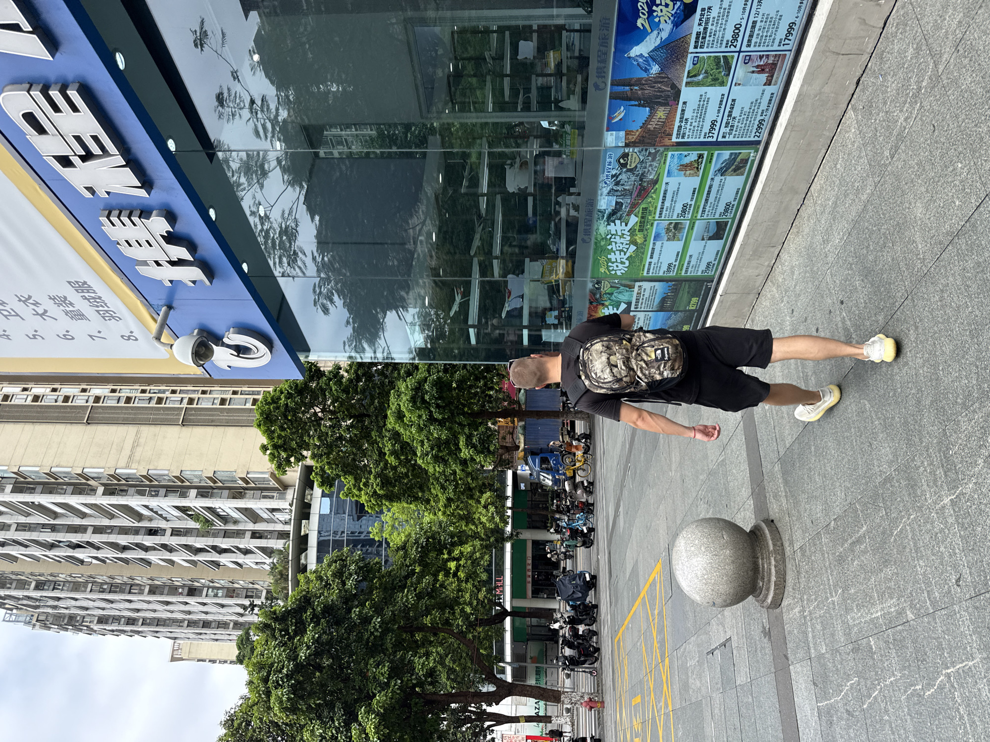 A man wearing a camouflage backpack and black shorts walks away from the camera on a paved city sidewalk in Tianhe District, China. He is wearing white sneakers. He is walking alongside a building with a blue facade and large glass windows displaying travel advertisements. The advertisements feature images of various tourist destinations and prices. Trees with green foliage are visible to the left of the man and the building. In the background, a tall, multi-story building with many windows and balconies dominates the skyline. Several parked scooters and a blue truck are visible in the street area further down the sidewalk. A grey spherical bollard is positioned on the sidewalk to the left of the man. The sky is overcast.