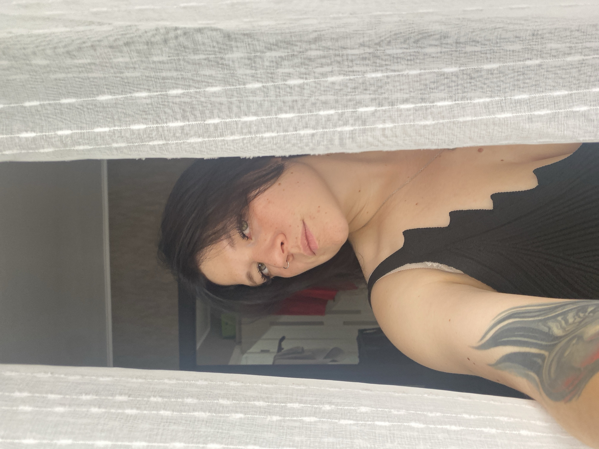 A young woman with dark hair, fair skin, and a nose ring is peeking through a white sheer curtain. She is wearing a black tank top and has a tattoo on her left arm. The background appears to be a room with a closet visible. The image was taken in Chaikovsky, Russia.