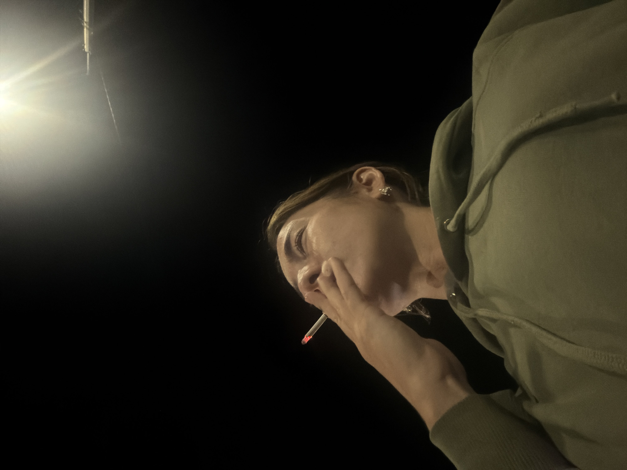 A low-angle medium shot captures a person smoking a cigarette at night. The person, viewed from the chest up, is looking upwards, with their face partially obscured by their hand holding the cigarette. The cigarette tip is glowing red, indicating it is lit. The person is wearing a green hooded sweatshirt. In the upper right corner of the image, a bright light source, possibly a streetlamp or overhead light, illuminates the dark sky, casting rays of light. The background is predominantly black, suggesting an outdoor urban environment at night. The specific location in Ust-Ilimsk, Russia, is not determinable from the visual information present in the image.