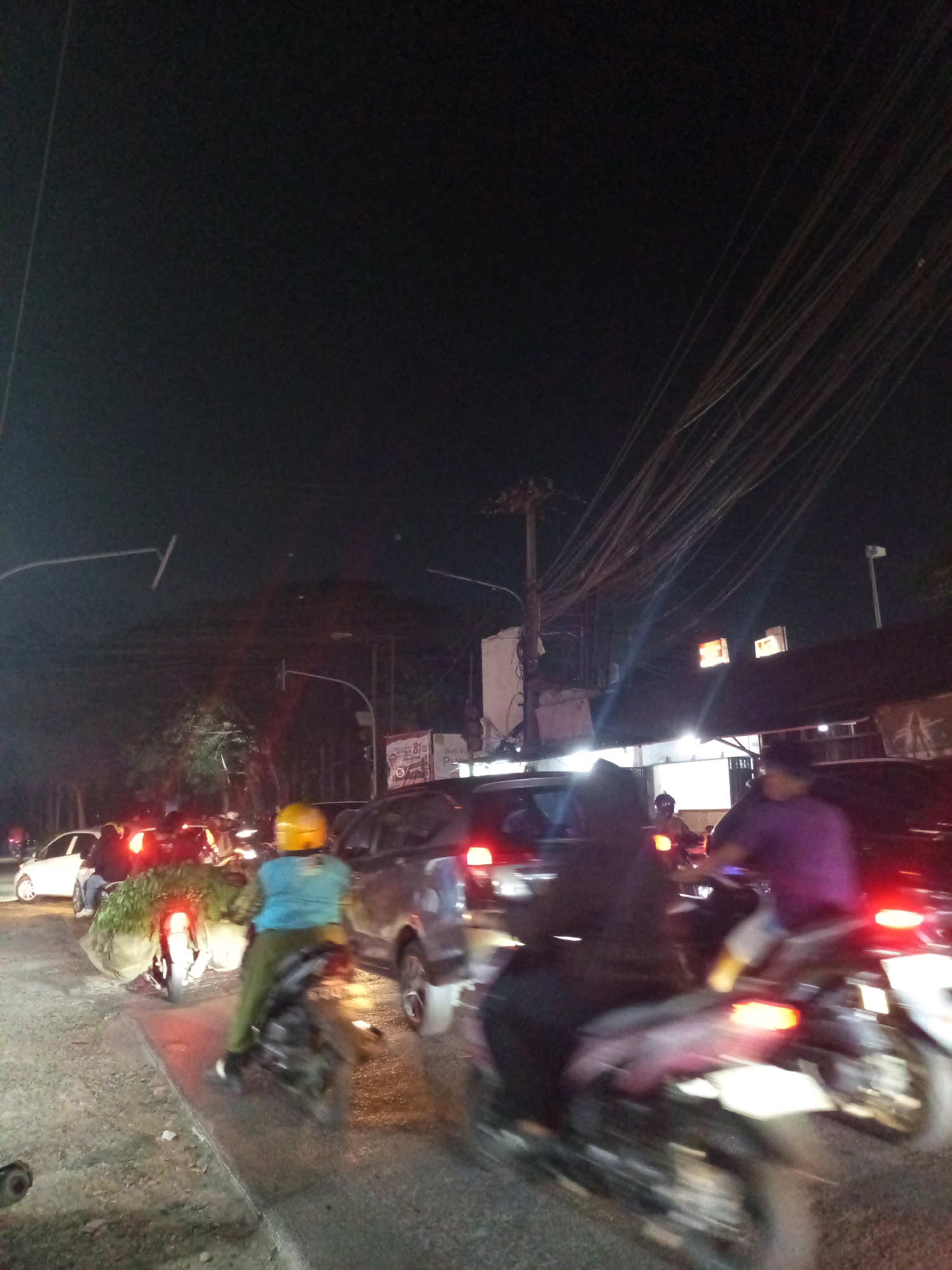 The image depicts a night scene in Karangsatria, Indonesia, showing a traffic jam. Several vehicles, including cars and motorcycles, are visible on a paved road. A motorcycle in the foreground has a rider wearing a blue shirt and a yellow helmet. Another motorcycle to the right is ridden by a person in a purple shirt. Buildings and streetlights line the road. Dense overhead wires are prominent in the upper portion of the image. The overall impression is one of congested urban nighttime traffic.
