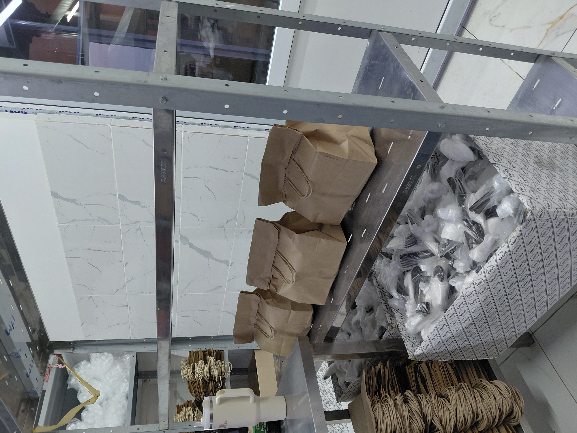 The image shows a metal shelving unit in what appears to be a commercial kitchen or storage area. On the shelves are stacks of brown paper bags, a large white thermal container, and a box filled with individually wrapped cutlery sets. The walls are tiled with a marble-like pattern. The scene is indoors and appears to be well-lit. There are no people visible in the image. The presence of food packaging suggests a food service establishment. The specific location in Aktau, Kazakhstan, cannot be determined from the visual information provided.