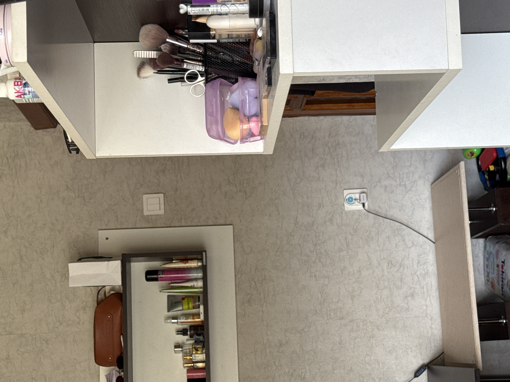 The image shows a dimly lit room, possibly a bedroom or dressing area, with a wall textured like rough plaster. On the left, a narrow shelf is mounted on the wall, holding various cosmetic items, including bottles, tubes, and a brown clutch bag. Above this shelf, a white paper bag is visible. To the right, a white shelving unit is present, with several shelves stacked vertically. The top shelf of this unit displays makeup brushes in a black container, a purple plastic case with makeup sponges, and other cosmetic products. A light switch is visible on the wall to the left of the shelving unit. Below the shelves, a thin wooden surface extends, supported by unseen structures. A power outlet with a blue circular element is mounted on the wall below the wooden surface, with a black wire extending from it. In the lower right of the image, the corner of a white furniture piece is visible. The overall impression is of a cluttered personal space, likely containing items related to makeup and personal care. The mention of "Яромінскі сельскі Савет, Belarus" is external information and not visually present in the image.