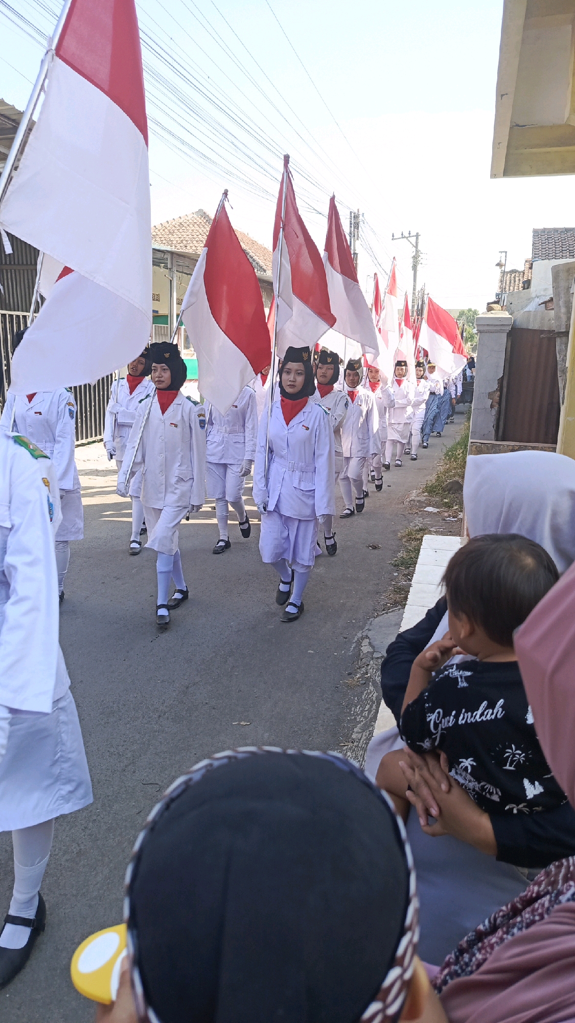 In the city of Pekauman Kulon, Indonesia, a group of young people in white uniforms with red scarves are marching in a parade, holding Indonesian flags. They are dressed in formal attire, complete with black hats and shoes. The parade is taking place on a paved road, with houses and buildings lining the sides. In the foreground, a few individuals are observing the parade, with one person holding a yellow and white object. The sky is clear and bright, indicating a sunny day. The scene captures a patriotic event, likely a celebration of Indonesian independence.