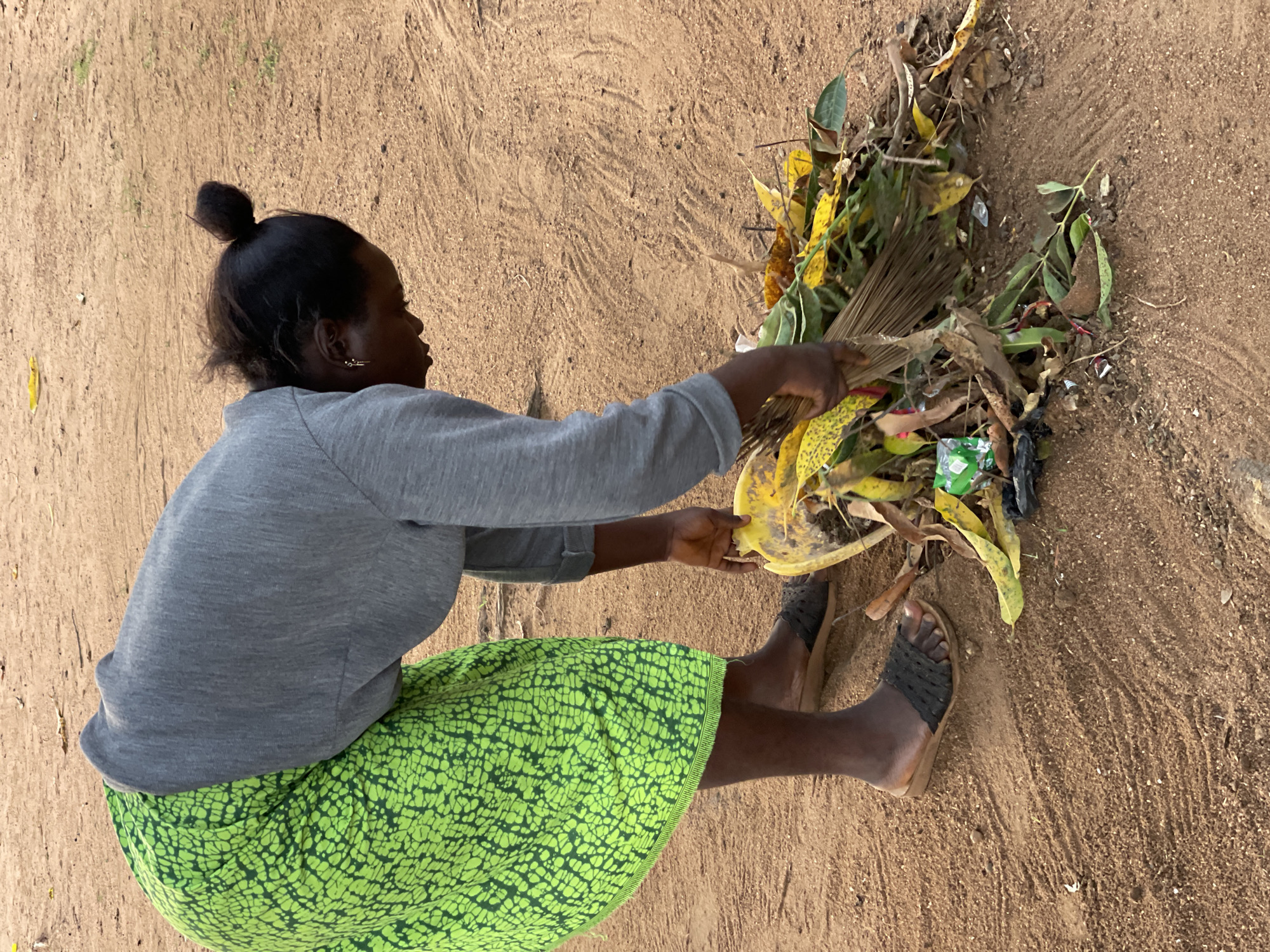 A woman is sweeping up leaves and trash on a dirt ground in Andaha, Nigeria. She is wearing a gray long-sleeved shirt, a green patterned skirt, and sandals. Her hair is pulled back in a bun. She is using a broom made of sticks to gather the debris into a pile. The pile contains leaves, twigs, and some plastic trash. The ground is dry and dusty.