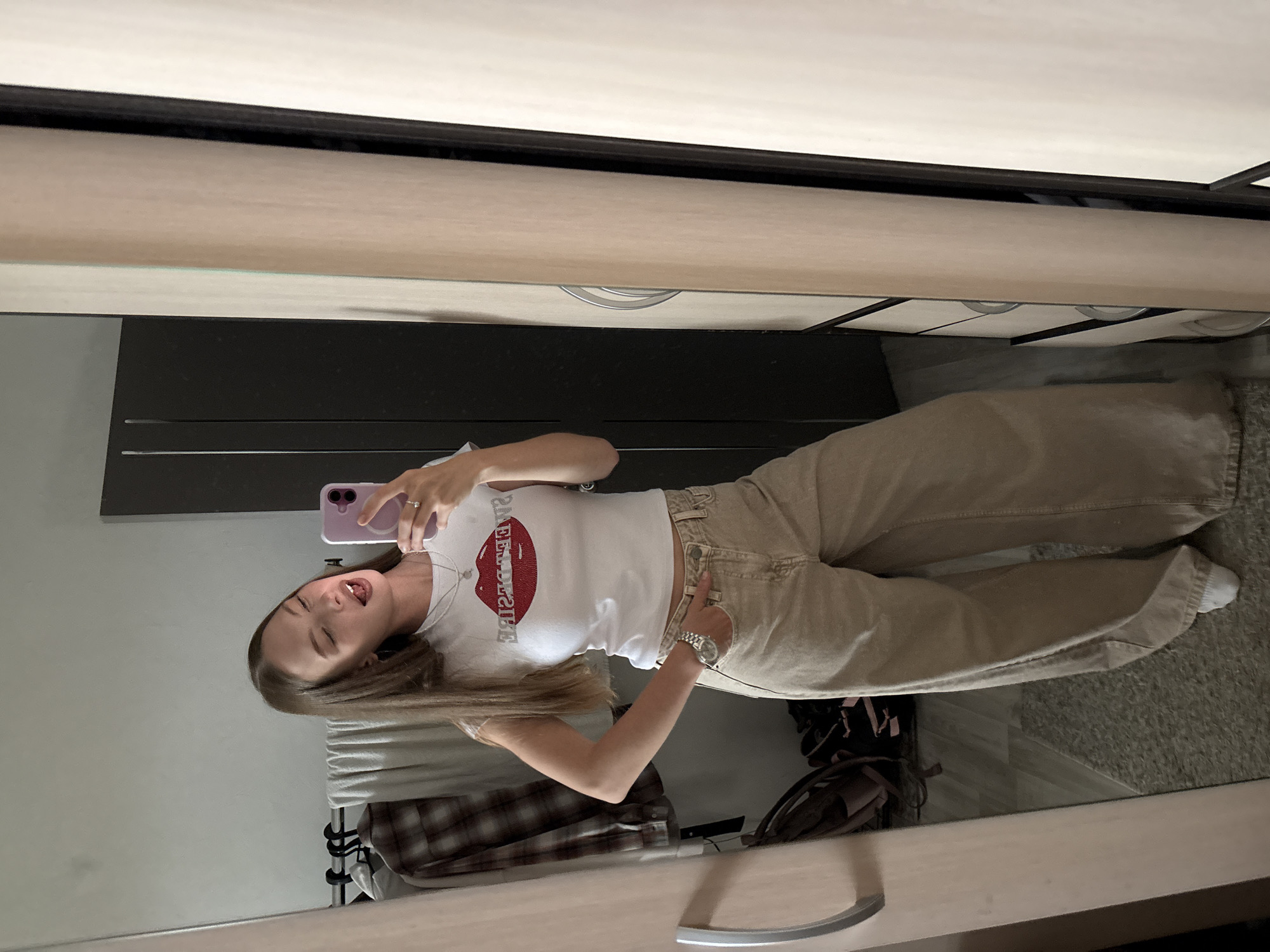 A young adult female, with long, straight, light brown hair and a fair complexion, takes a full-body mirror selfie in what appears to be a domestic interior in Perm, Russia. She is wearing a white, short-sleeved, cropped t-shirt featuring a prominent red lip graphic with the mirrored text "SWEET DESIRE" across the chest. Her lower attire consists of light beige or khaki wide-leg jeans, and white socks are visible at her ankles.
She holds a lavender-colored smartphone in her left hand, which is raised to capture her reflection in the full-length mirror. Her right hand is positioned on her hip. Her facial expression is playful, characterized by a slight wink or partially closed right eye and an open mouth revealing a portion of her tongue. Accessories include a silver-toned watch on her left wrist, a simple silver-colored necklace with a small pendant, and a ring on her right ring finger.
The mirror reflects the room behind her. To the left of the subject, a clothing rack displays several items, including a brown plaid shirt, a white t-shirt, and a white towel. Below the rack, on a light grey speckled rug or carpet, a dark-colored backpack with pink accents is visible, alongside other dark items, possibly shoes or another bag. A light grey textured wall is visible behind the clothing rack, with portions of dark gray furniture or a door featuring subtle vertical grooves further in the background. To the right of the subject, the reflection shows the edge of a light-colored wooden dresser or closet with white drawers and silver circular pull handles. A curved silver handle is also reflected on a door or panel further to the right. The overall lighting is bright and even, indicating an indoor setting likely illuminated by ambient light.
