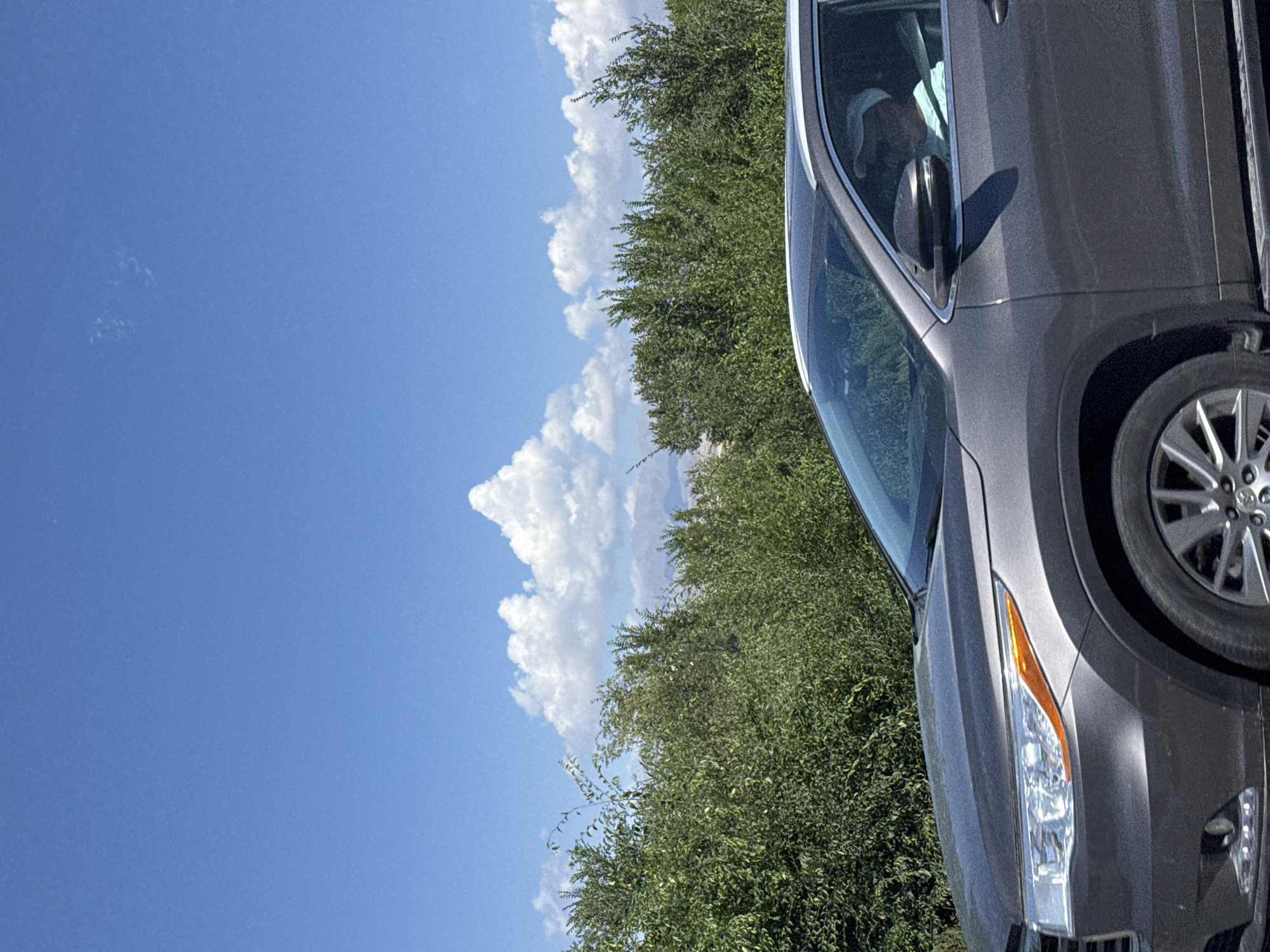 The image captures a dark gray SUV driving on a road with lush green foliage lining the roadside. The sky above is a vibrant blue, dotted with fluffy white clouds. In the distance, hazy mountains can be seen against the horizon. A person wearing a white cap and a light blue shirt is visible inside the SUV, seated in the driver's seat. The scene appears to be set in Toru-Aigyr, Kyrgyzstan, with a natural, outdoor environment.
