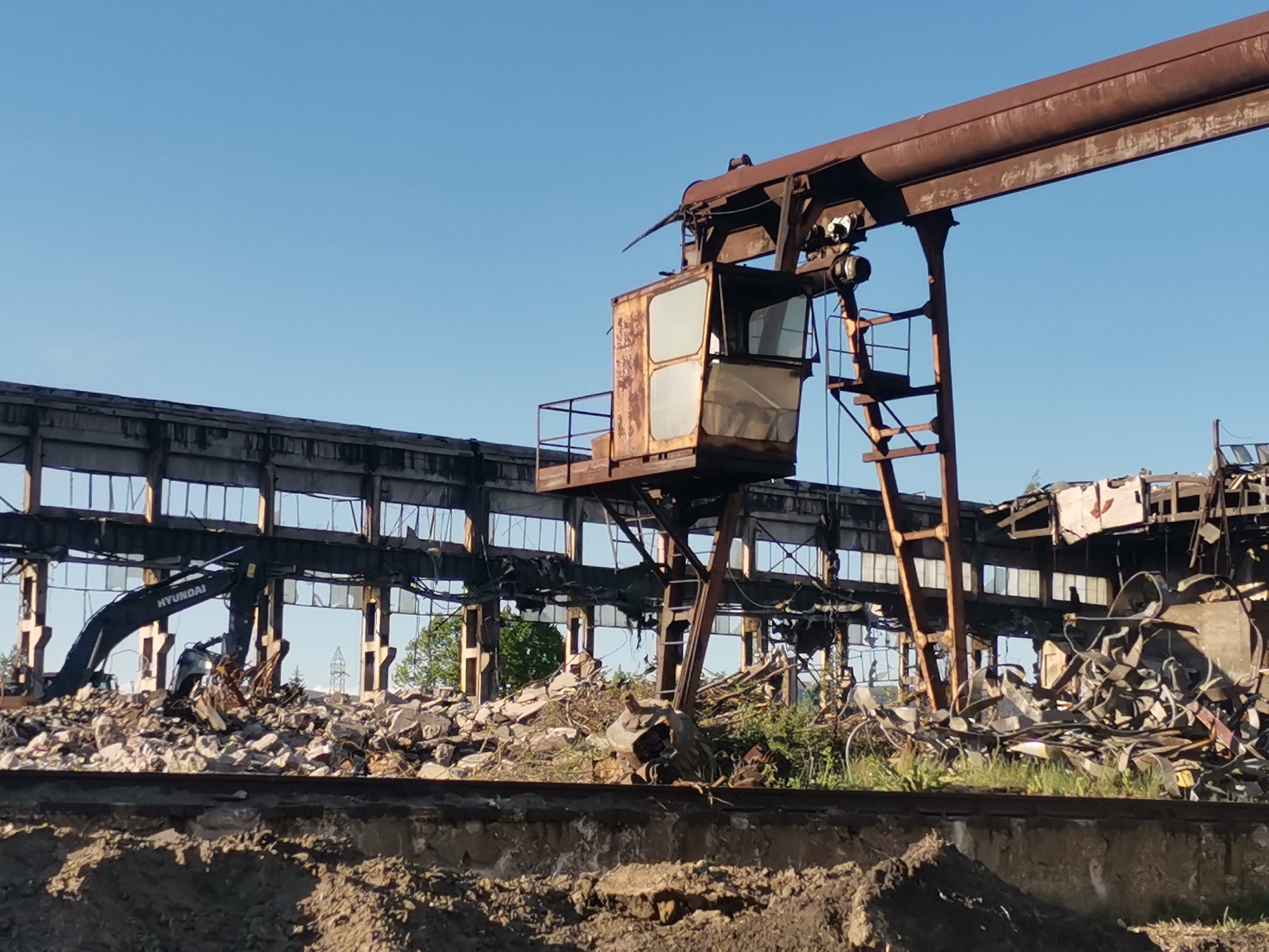 The image depicts the remnants of an industrial site, likely an abandoned factory, in Zhivkovo, Bulgaria. A large overhead crane with a rusted control cabin dominates the foreground. In the background, a partially destroyed building frame stands amidst piles of rubble and debris. A Hyundai excavator is visible, indicating ongoing demolition or cleanup activity. The ground is uneven and covered in dirt and vegetation. A set of railroad tracks runs parallel to the debris pile in the middle ground. The sky is clear and blue, suggesting a bright, sunny day.