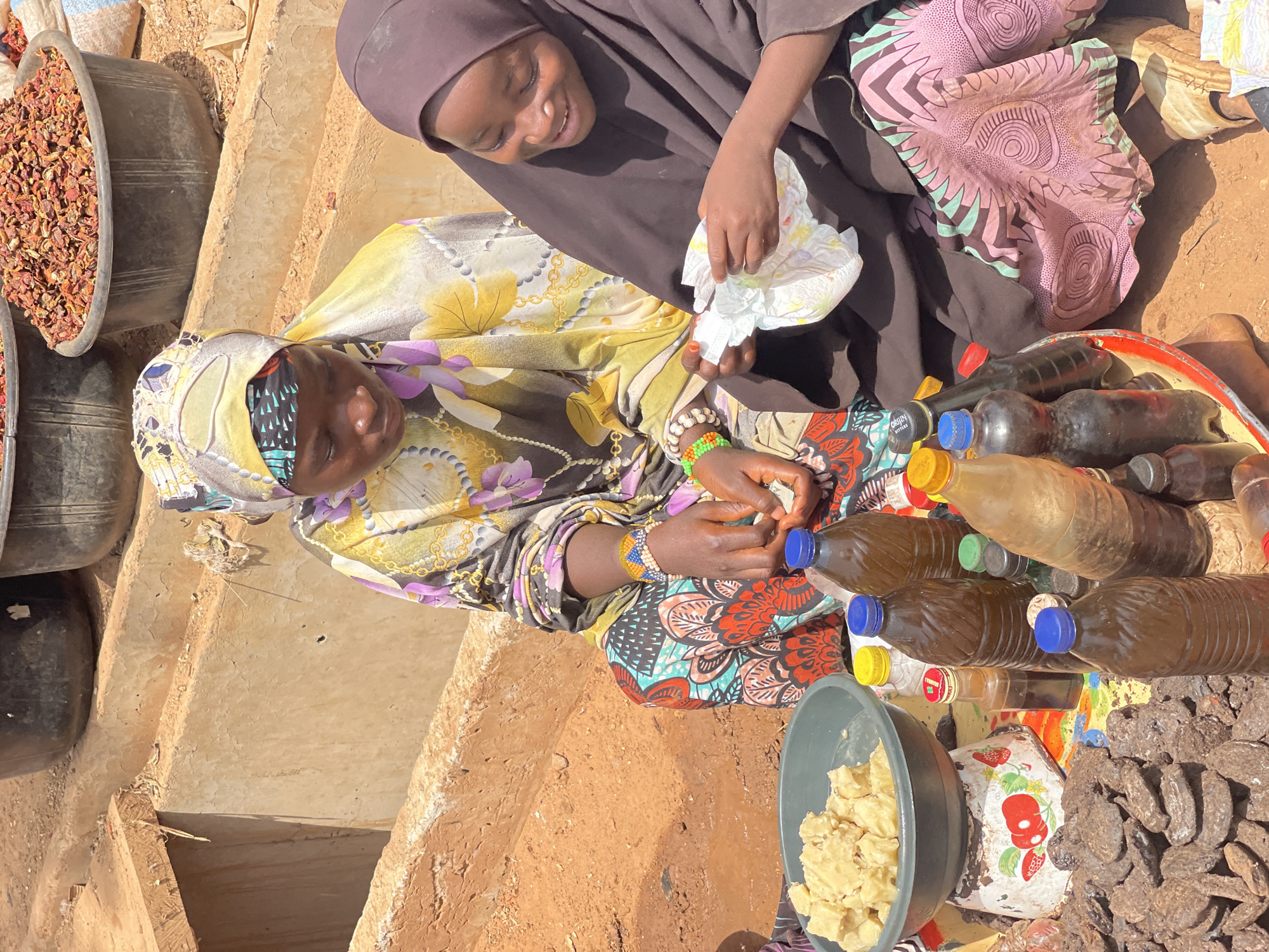 Two females are seated on a weathered, earth-toned platform, likely at a marketplace in Zaria, Nigeria. The female on the left wears a floral-patterned yellow and purple headscarf and blouse over a colorful patterned skirt. She is facing the right side of the scene and working with her hands. On the right, a younger female wearing a dark brown hijab and floral print garment looks down, manipulating a white object in her hands. A tray containing numerous plastic bottles of dark liquid is positioned in front of the women, with a grey bowl of light-colored chunks and a pile of dark, dried objects nearby. Two black tubs filled with dried reddish material rest on a higher tier to the left. The overall environment appears to be an outdoor marketplace on a sunny day.