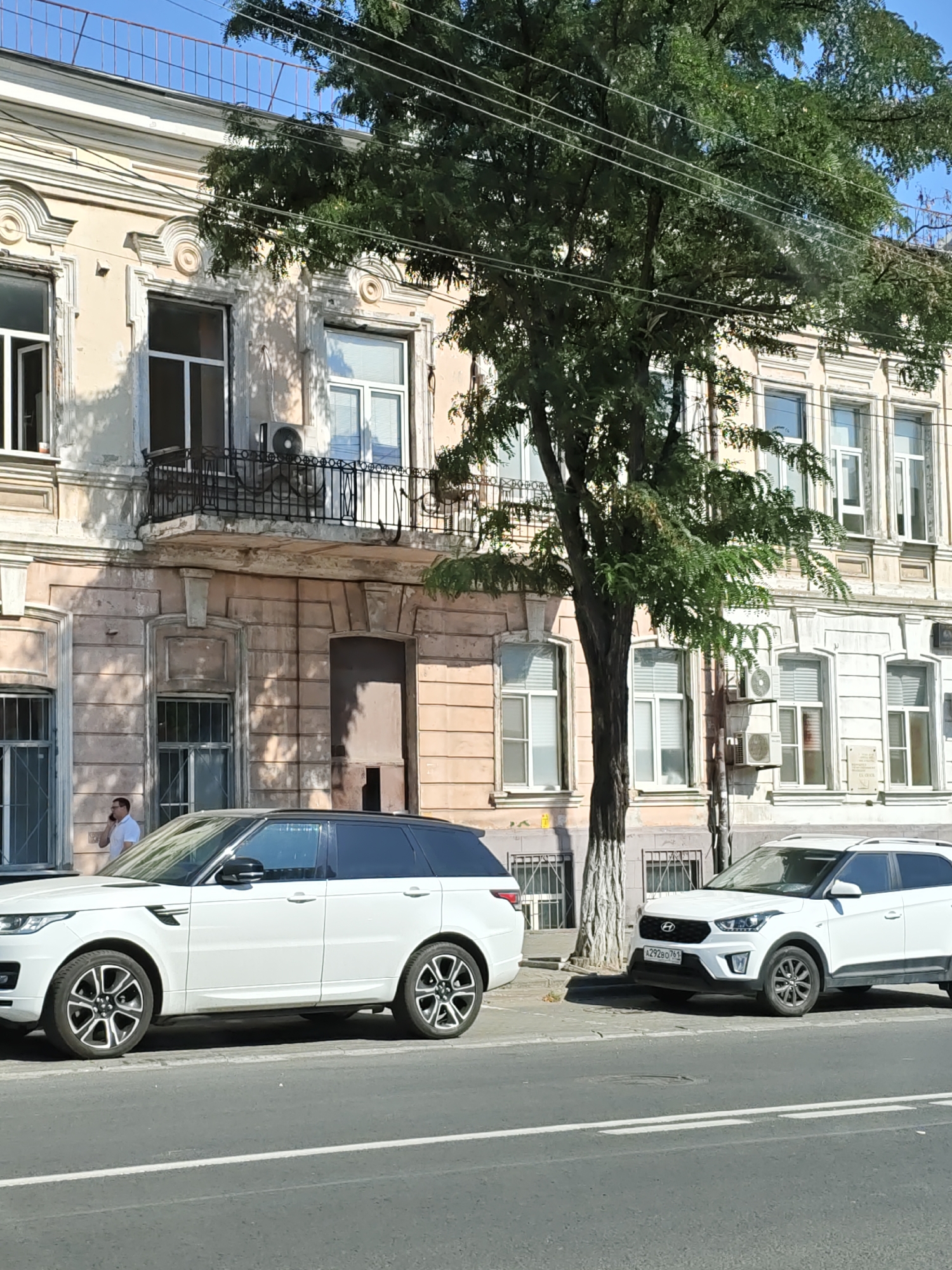 A white Range Rover Sport is parked on the street in front of an old building in Rostov-on-Don, Russia. A white Hyundai Creta is parked to the right of the Range Rover. A man in a white shirt is standing on the sidewalk in front of the building, talking on his phone. The building appears to be old and somewhat run-down, with peeling paint and ornate architectural details. A large tree with green leaves is in front of the building, partially obscuring the view of the windows. The sky is clear and blue.
