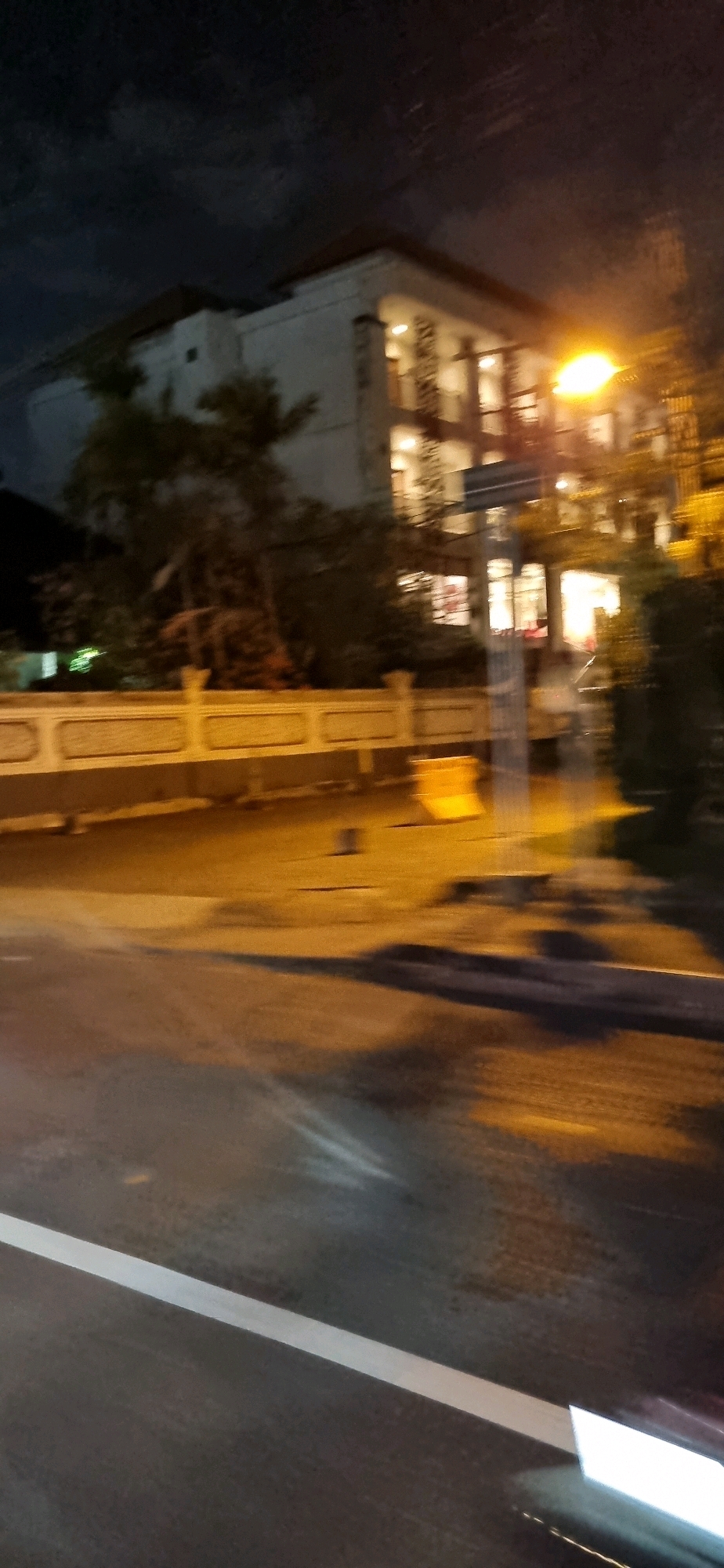 The image captures a nighttime scene in Denpasar, Indonesia, showing a multi-story building with illuminated windows. A street lamp with a bright orange glow is visible to the right of the building, casting light on the surroundings. In front of the building, a low, light-colored wall with decorative patterns runs along the street. The street itself is wet, reflecting the lights, and a white line marking the lane is visible on the asphalt. The overall impression is one of a blurred, possibly fast-moving shot taken at night, with artificial lighting dominating the scene.