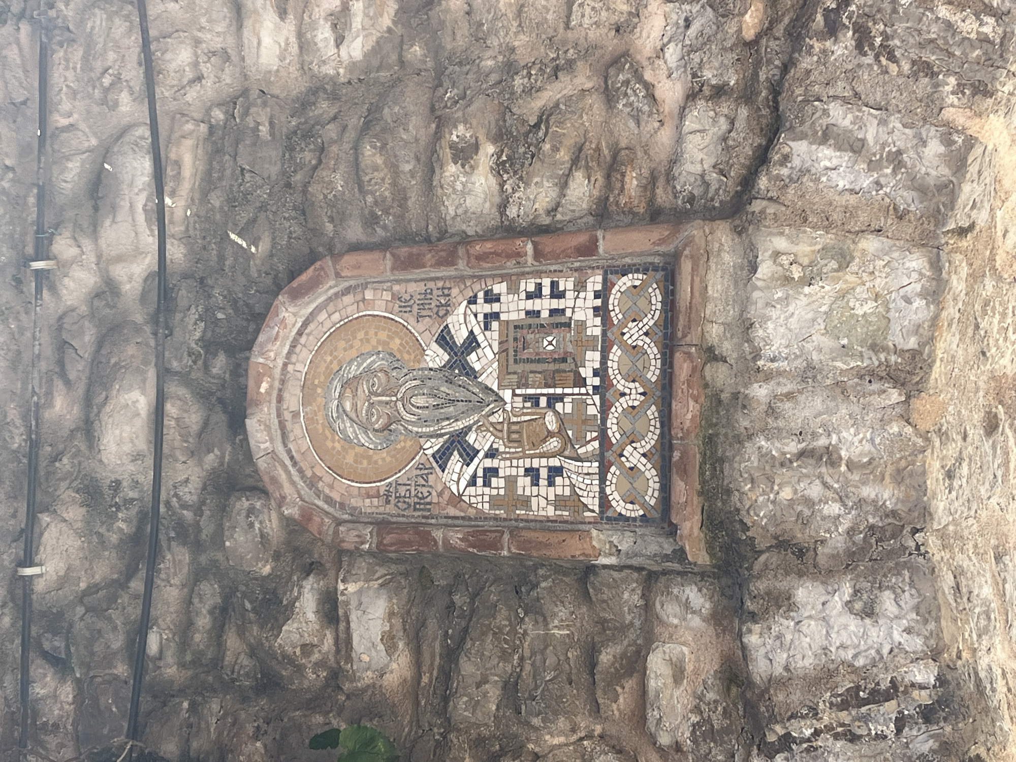 A mosaic icon of Saint Peter is embedded in a rough stone wall. The mosaic is arched at the top and depicts a bearded man with a halo, wearing patterned robes and holding a book and a staff. Cyrillic text is visible on either side of the saint's head. The stone wall is textured and weathered, with visible cracks and variations in color. There are some cables running across the top of the wall. The mosaic is located next to a stone archway, suggesting it is part of a religious site or building in Budva, Montenegro.