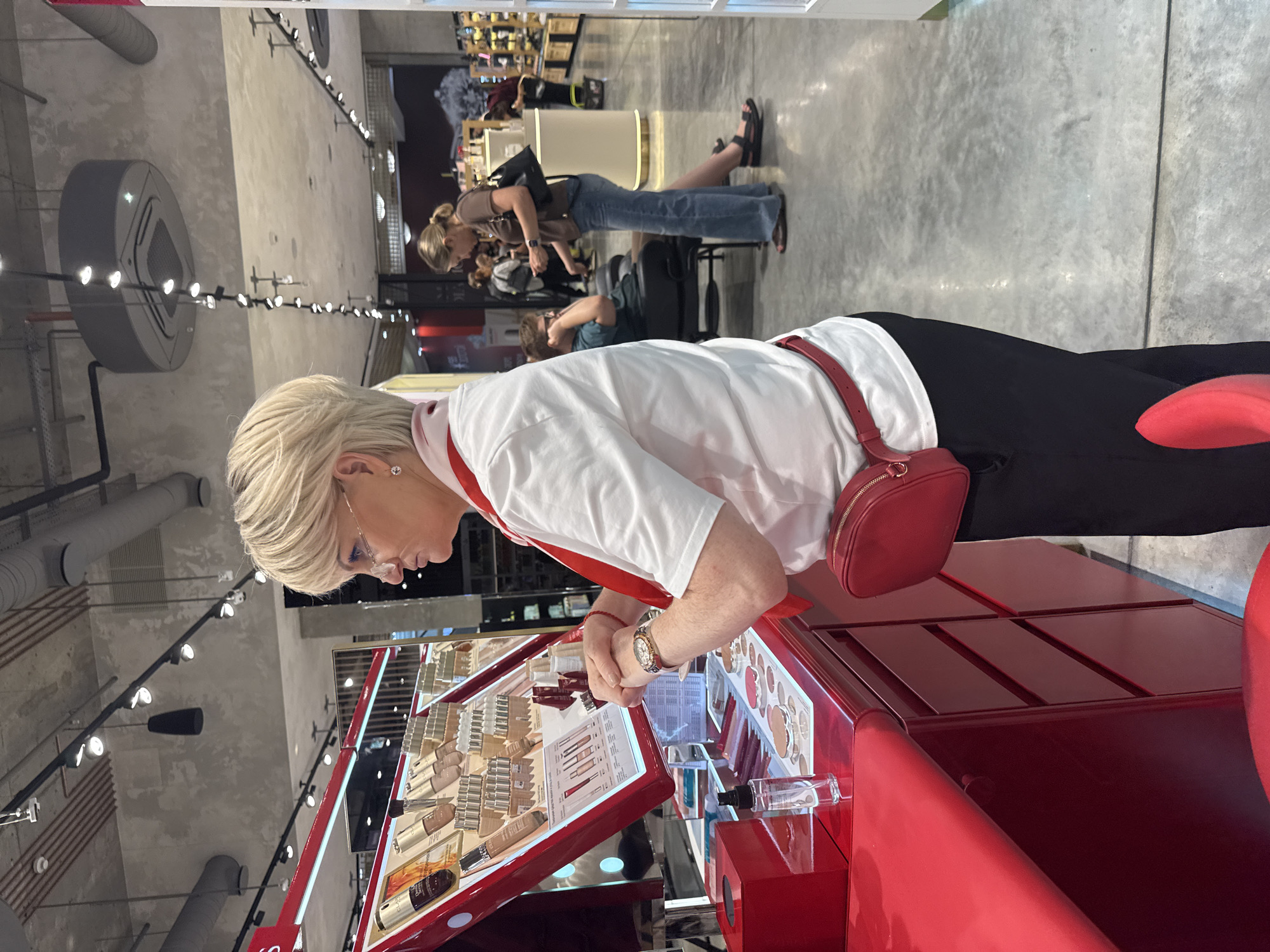 In Moscow, Russia, a woman with blonde hair, wearing glasses, a white shirt, black pants, and a red fanny pack, stands at a red counter displaying makeup products. She is looking at the products. In the background, other shoppers are visible in a retail store with a concrete floor and ceiling with exposed pipes and track lighting.