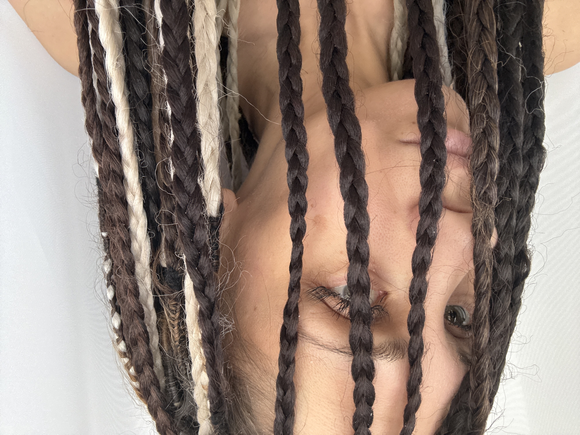Here is a forensic description of the image:
A person with braided hair is seen from an upside-down perspective. The braids are a mix of brown, black, and white/blonde colors. The braids are hanging down, partially obscuring the person's face. The skin tone of the person appears light. The person's eyes are visible through the gaps in the braids. The background appears to be a plain white surface. There is no information in the image regarding location of this scene.