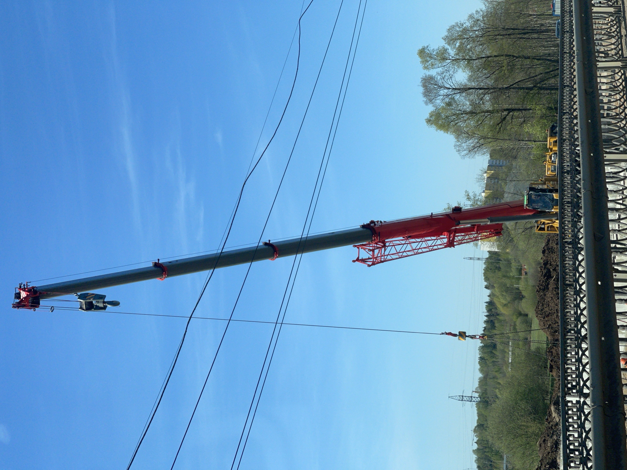The image depicts a large red and gray crane extending its boom across a landscape in Kabanikha, Russia. The crane is positioned next to a patterned metal fence, with trees and a distant building visible in the background. Power lines run across the sky above the crane's boom. Yellow construction equipment can be seen further back, near the base of the trees. The sky is bright blue with a few wispy clouds.