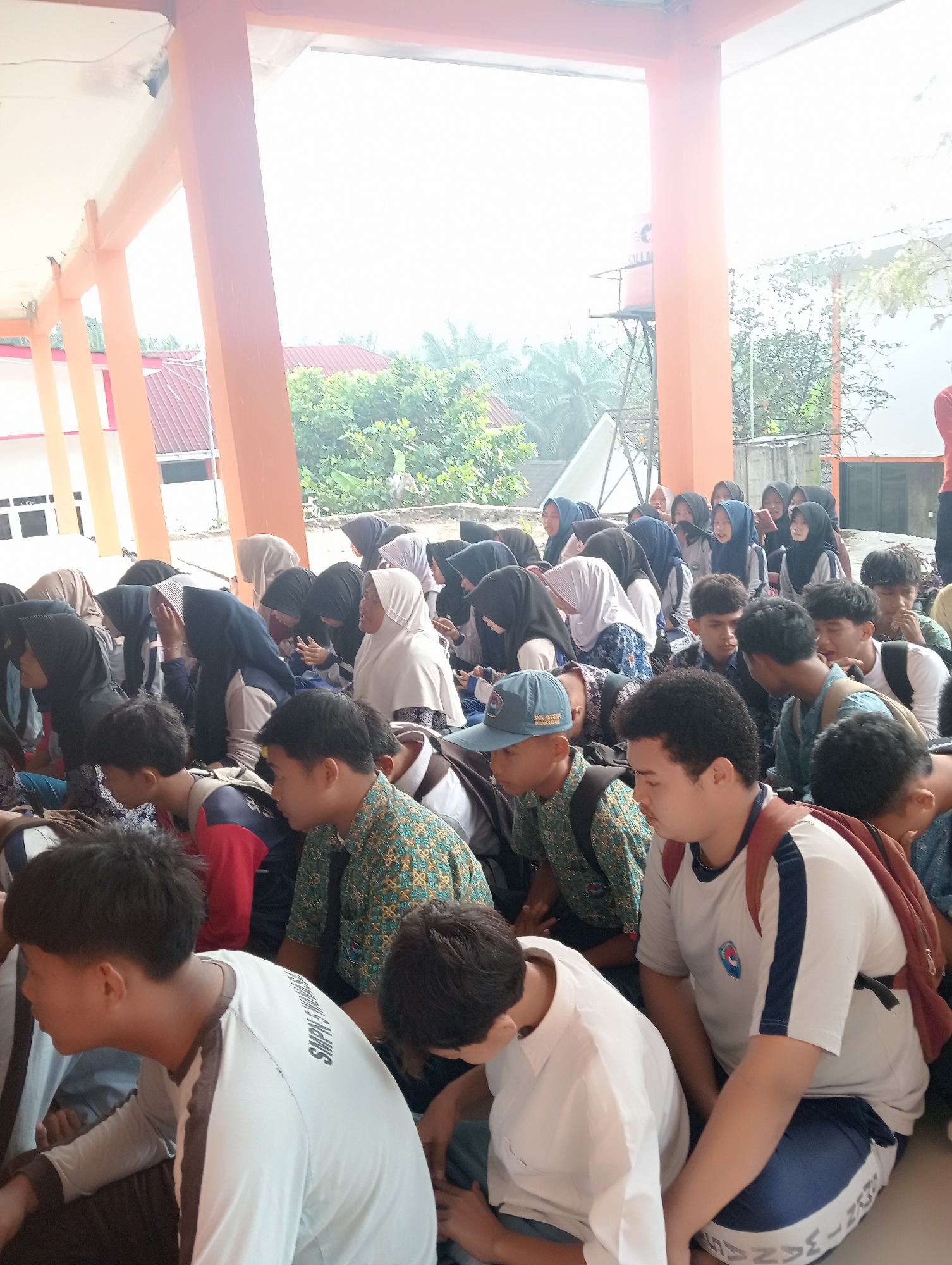 The image depicts a group of students, primarily young males and females, sitting on the ground in an outdoor covered area. The architecture suggests it is likely a school or community building with orange pillars and a white ceiling. Many of the females are wearing hijabs. The students appear to be engaged in some activity, possibly a gathering or lesson, as they are all facing in roughly the same direction. In the background, lush green trees and parts of buildings are visible, indicating a tropical or subtropical environment. The presence of a water tower in the background further suggests an outdoor setting. The overall scene is informal and suggests a communal activity within a school context, likely in Wanasalam, Indonesia, given the style of dress and surrounding vegetation.