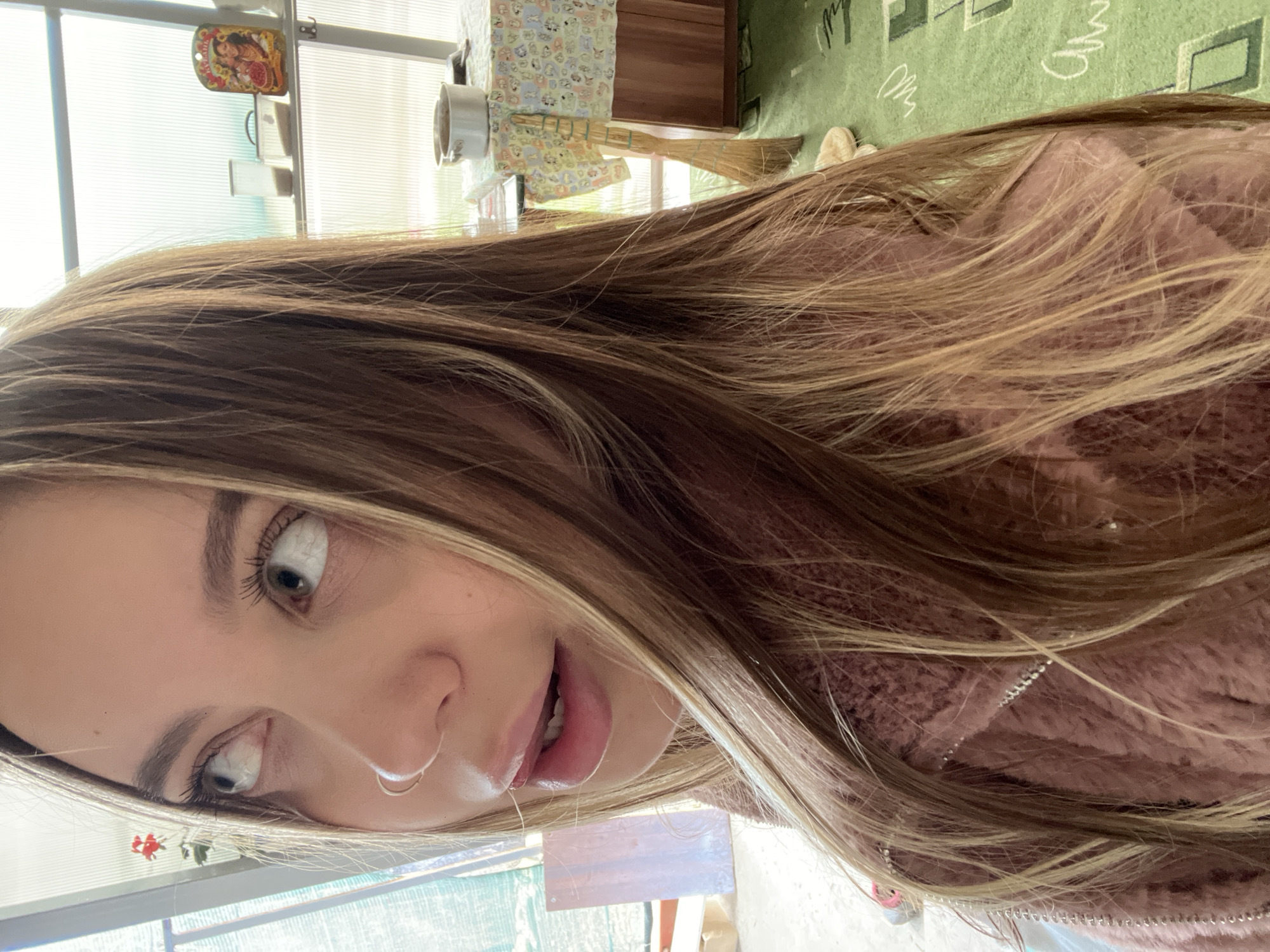 A close-up, angled selfie of a woman with light brown hair and light eyes. She has a nose ring and is wearing a pink, fuzzy jacket. The background shows a room with a window, part of a table covered with a patterned cloth, and a section of a green rug. The location is Novopokrovskaya, Russia. The context of the image does not provide any further detail.