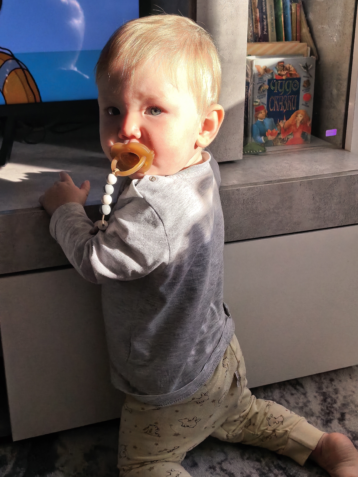 A toddler with light blonde hair and blue eyes is captured kneeling on a patterned grey rug, looking directly towards the viewer. The child has a translucent brown pacifier in its mouth, attached by a white and light blue beaded chain to a grey long-sleeved shirt with two visible snap buttons at the collar. The toddler is wearing light yellow pants adorned with repetitive brown cartoon animal (likely rabbit) patterns. The child's right hand grasps the edge of a grey, textured, multi-level piece of furniture that serves as a media console or shelving unit. To the left, a television screen displays a cartoon character with orange and white stripes against a blue background, with a distinct bright, cloud-like reflection. To the right, within a compartment of the furniture, several books are visible; the most prominent cover is titled "ЧУДО СКАЗКИ" (Wonder Tales) in Cyrillic script, depicting a boy in blue holding a red object and a girl in red. Strong natural light originates from the left side of the frame, creating defined shadows on the furniture and the toddler's clothing. This scene is located in the city of Sterlitamak, Russia.