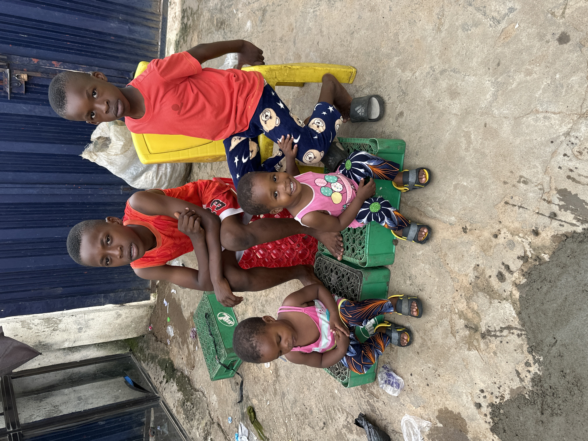 In Narayi, Nigeria, four young children, two boys and two girls, are seated outdoors on a concrete surface. The two older children, boys, are positioned behind the two younger girls. The boy on the left wears a red tank top and white shorts, while the boy on the right is dressed in a red t-shirt and patterned blue pants. The younger girls are seated on green plastic crates. The girl on the left wears a pink and white striped tank top and patterned pants. The girl on the right is in a pink tank top adorned with cartoon balloons and matching patterned pants. The older boy on the right is seated on a yellow plastic chair. The background features a blue corrugated metal fence and a rough concrete wall with a broken window. Various items are scattered on the ground, including plastic bags, debris, and what appears to be fresh cement.