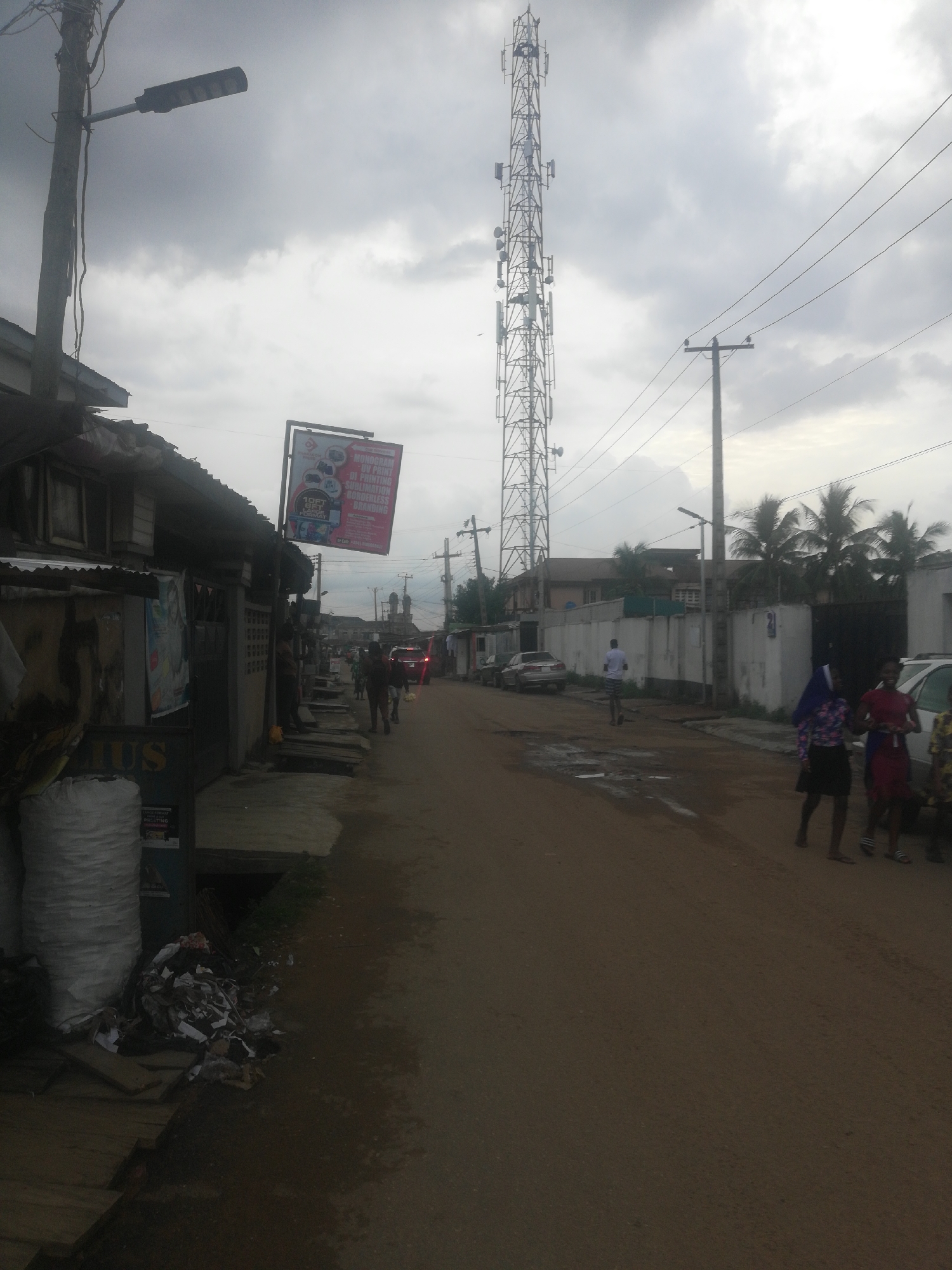 The image depicts a street in Lagos, Nigeria on an overcast day. The road is unpaved and wet, with a large puddle in the middle.
The buildings on the left are dilapidated, with corrugated iron roofs and exposed wooden structures. There is a sign for a business on the left and piles of trash in front of the building.
A tall telecommunications tower dominates the skyline in the center of the image, and utility poles with power lines run along the right side of the street.
Several people are walking along the road, some of whom are women in traditional clothing. Cars are parked on the side of the road. The overall scene suggests a low-income residential area.