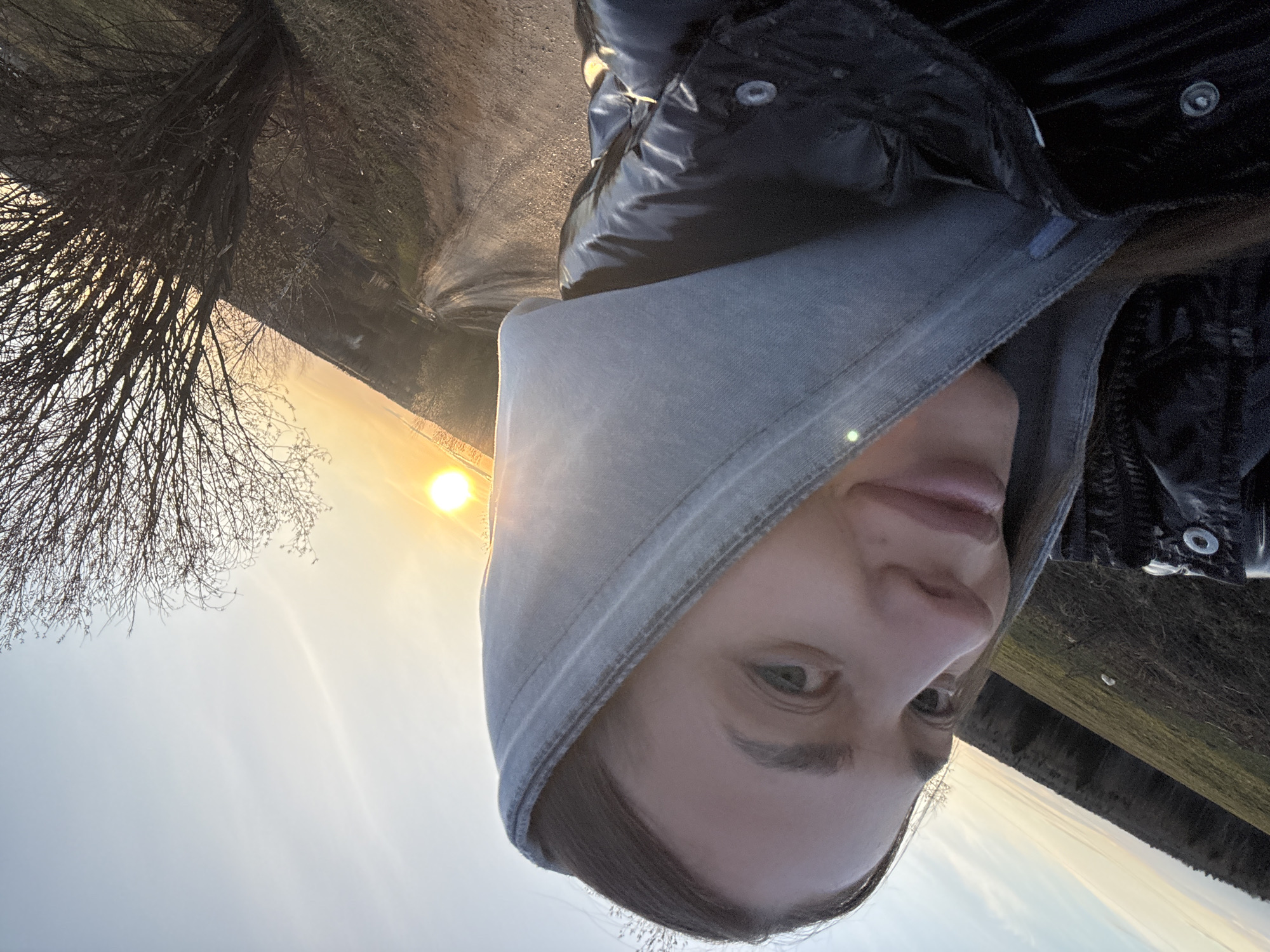 A woman wearing a gray hooded jacket and a black puffy coat is taking a selfie outdoors. The background shows a bare tree, a path, and the sun low in the sky. The environment appears to be rural or on the outskirts of a city, though there is no clear indication it is specifically in Amin'kai, Russia.