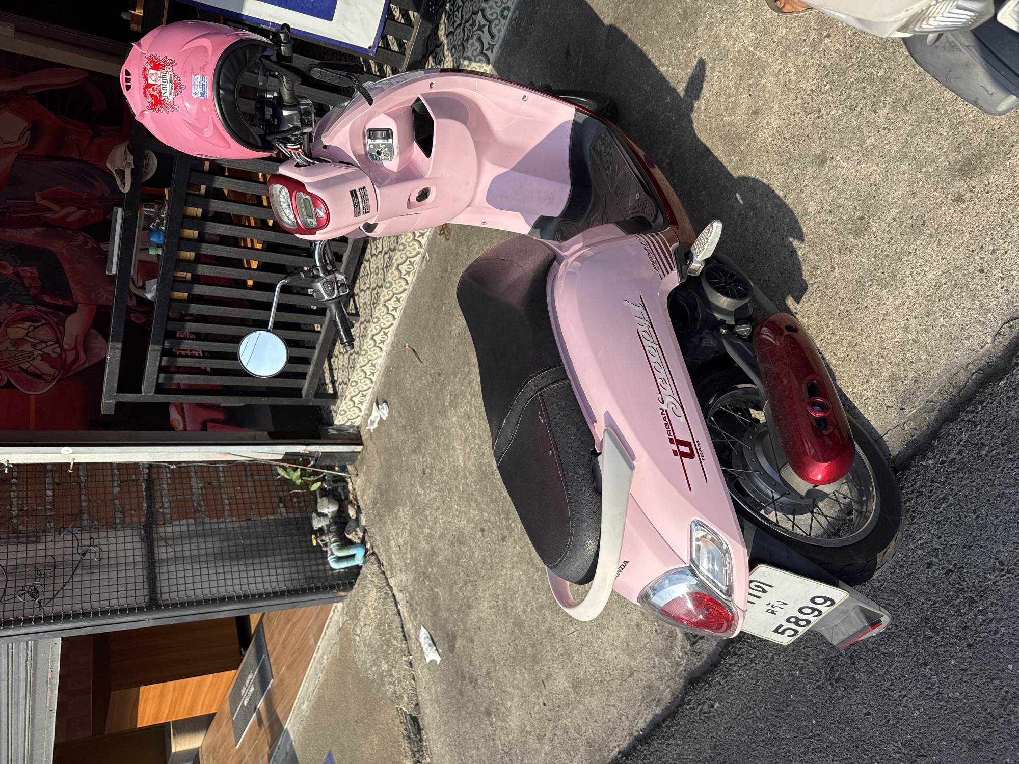 A pink Honda scooter with license plate number 5899 is parked on a concrete street in Phuket City Municipality, Thailand. The scooter has a black seat and is parked near a building with a black metal gate. A pink helmet with a graphic design is resting on the scooter's handlebars. A person is partially visible in the background.