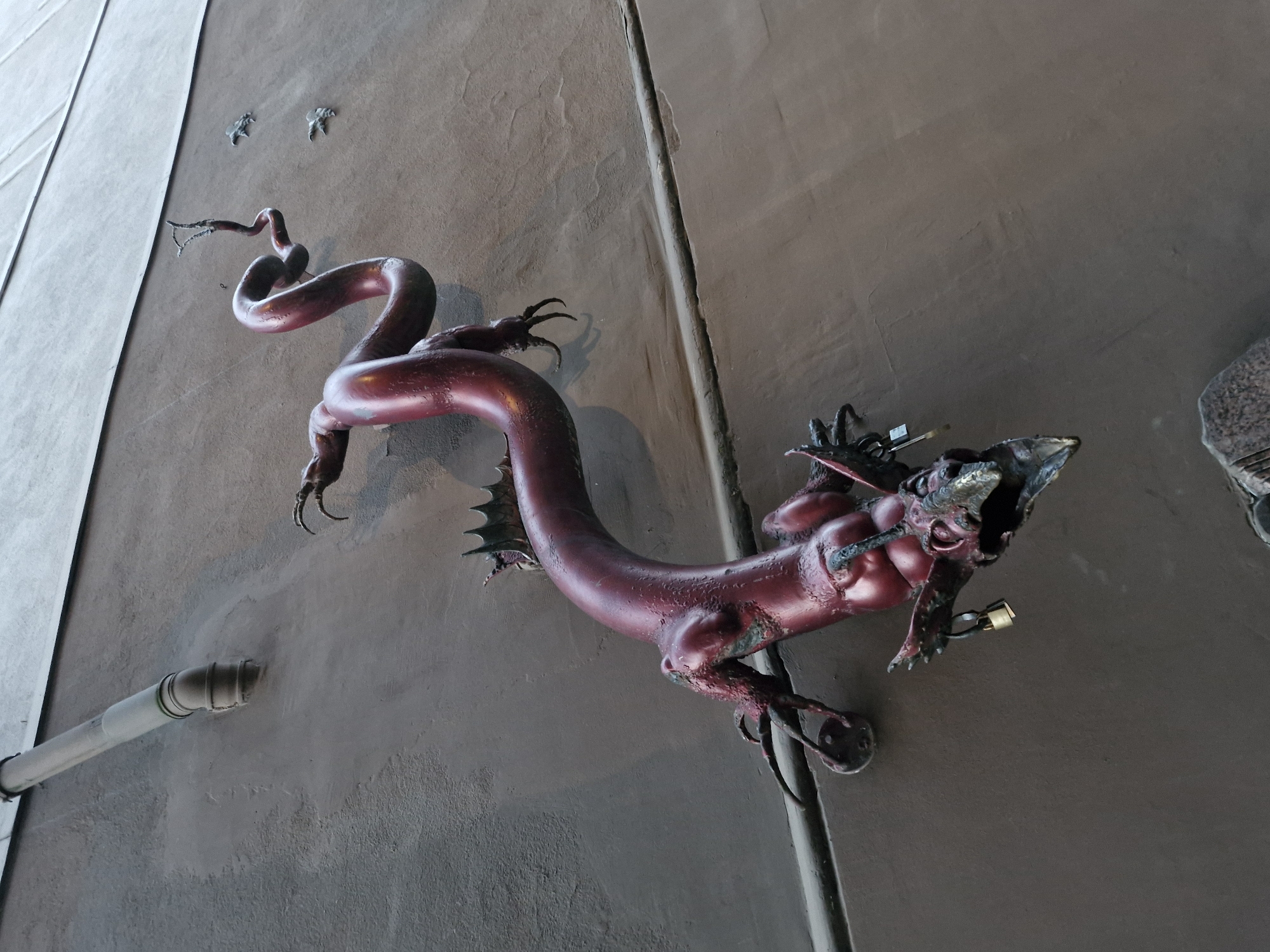 The image shows a dark red metallic sculpture of a dragon-like creature fixed to a brown building wall in Klaipėda, Lithuania. The sculpture depicts a sinuous, serpentine body with prominent ribs, a spiky dorsal fin, clawed limbs, and a detailed head with sharp teeth and a beak-like snout. A small padlock is attached to one of the creature's forelimbs. In the upper left corner of the image, a section of a light-colored pipe, likely a drainpipe, is visible against a lighter grey surface of the building. The building wall has a textured finish, and a horizontal line, possibly a joint or molding, runs across the wall. Two small, metallic dragon claw sculptures are mounted on the wall to the right of the main sculpture. The overall impression is of a whimsical or perhaps slightly menacing piece of public art.