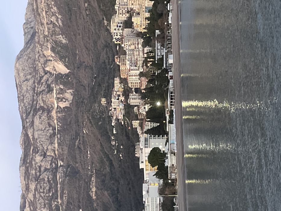 A towering, rocky mountain dominates the left side of the image, its rugged terrain catching the sunlight. Adjacent to the mountain, a cluster of buildings cascades down the hillside towards the coast. The structures, varying in size and color, create a dense urban landscape. A narrow strip of beach separates the buildings from the water. The sea, reflecting the sky, forms a gray expanse on the right, with faint light reflections shimmering on its surface.