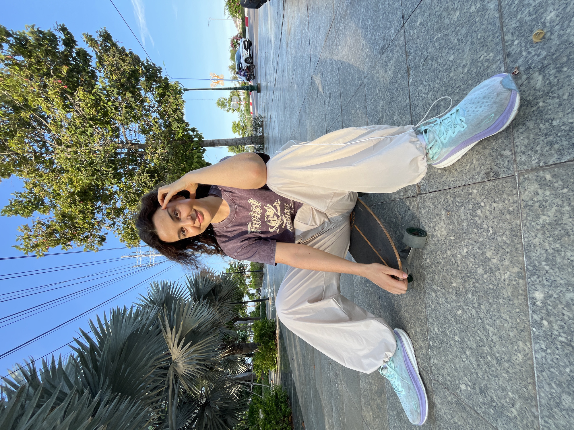 A young woman with curly brown hair sits on a skateboard on a paved walkway. She wears a purple t-shirt with a graphic print and loose, cream-colored pants. Her feet are clad in light blue and purple athletic shoes. The woman is positioned in the foreground, with her right hand resting on her forehead and her left hand near the skateboard. Behind her, palm trees and a leafy tree are visible against a clear blue sky. In the background, a street with a car and a motorbike can be seen. The setting appears to be an urban park or promenade in Phu Thuy Ward, Vietnam, indicated by the tropical vegetation and overhead power lines. The time of day is likely late afternoon, given the warm lighting and long shadows.