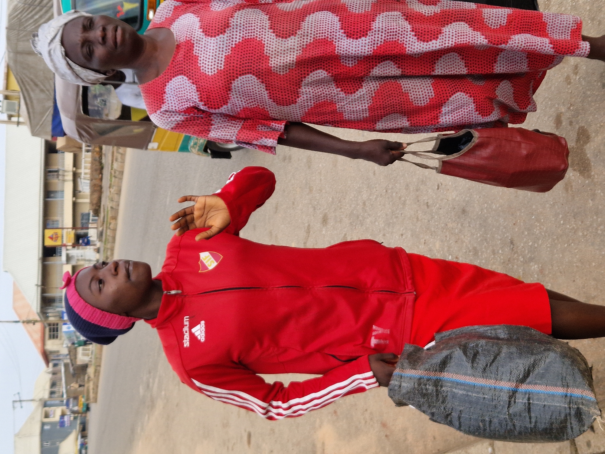 Two women are walking on a paved street in Kabong, Nigeria. The woman on the left is wearing a red tracksuit with white stripes on the sleeves, a blue and pink beanie, and a red skirt. She is carrying a large black bag in her left hand. The woman on the right is wearing a red and white patterned dress and a white headscarf. She is carrying a reddish-brown bag in her right hand. In the background, there is a yellow and green motorized tricycle, and buildings can be seen on either side of the street. The sky is overcast.