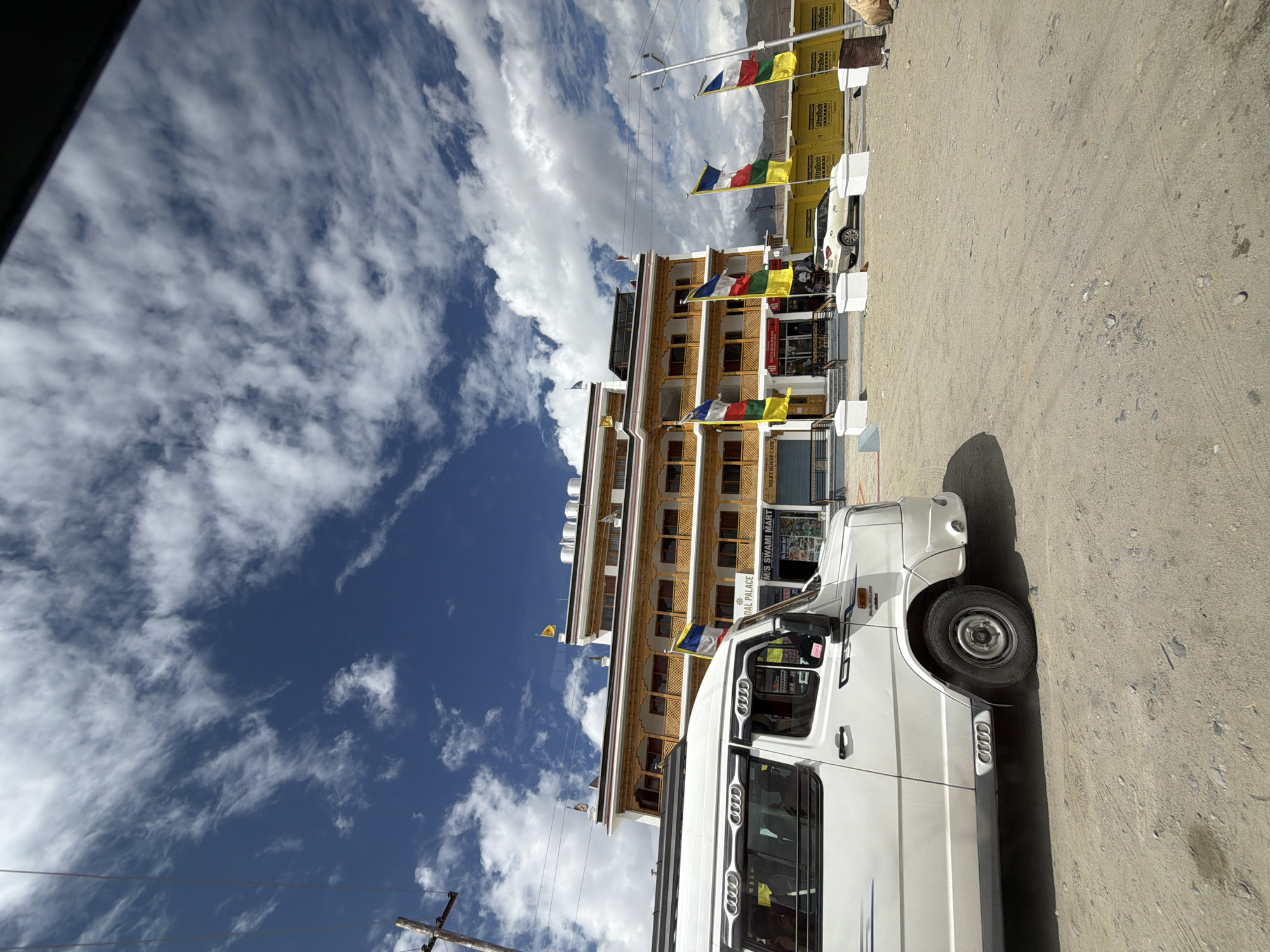A low-angle, full shot captures a white passenger van parked on a dusty road in front of a multi-story building with yellow trim. The van, a Force Traveller, is positioned on the left side of the frame, with its front right tire visible. Behind the van, the building, identified by signage as "OM PALACE," exhibits traditional Tibetan architecture with wooden balconies and prayer flags. Several flags in various colors, including red, yellow, blue, and white, are displayed throughout the scene. A white car is parked in front of the building, near a row of yellow barricades with the "Ultratech" logo. The sky above is a vibrant blue, filled with scattered white clouds. The ground is covered in dry, dusty soil, with scattered pebbles and a few dark stains. The environment suggests a remote, possibly mountainous, location.