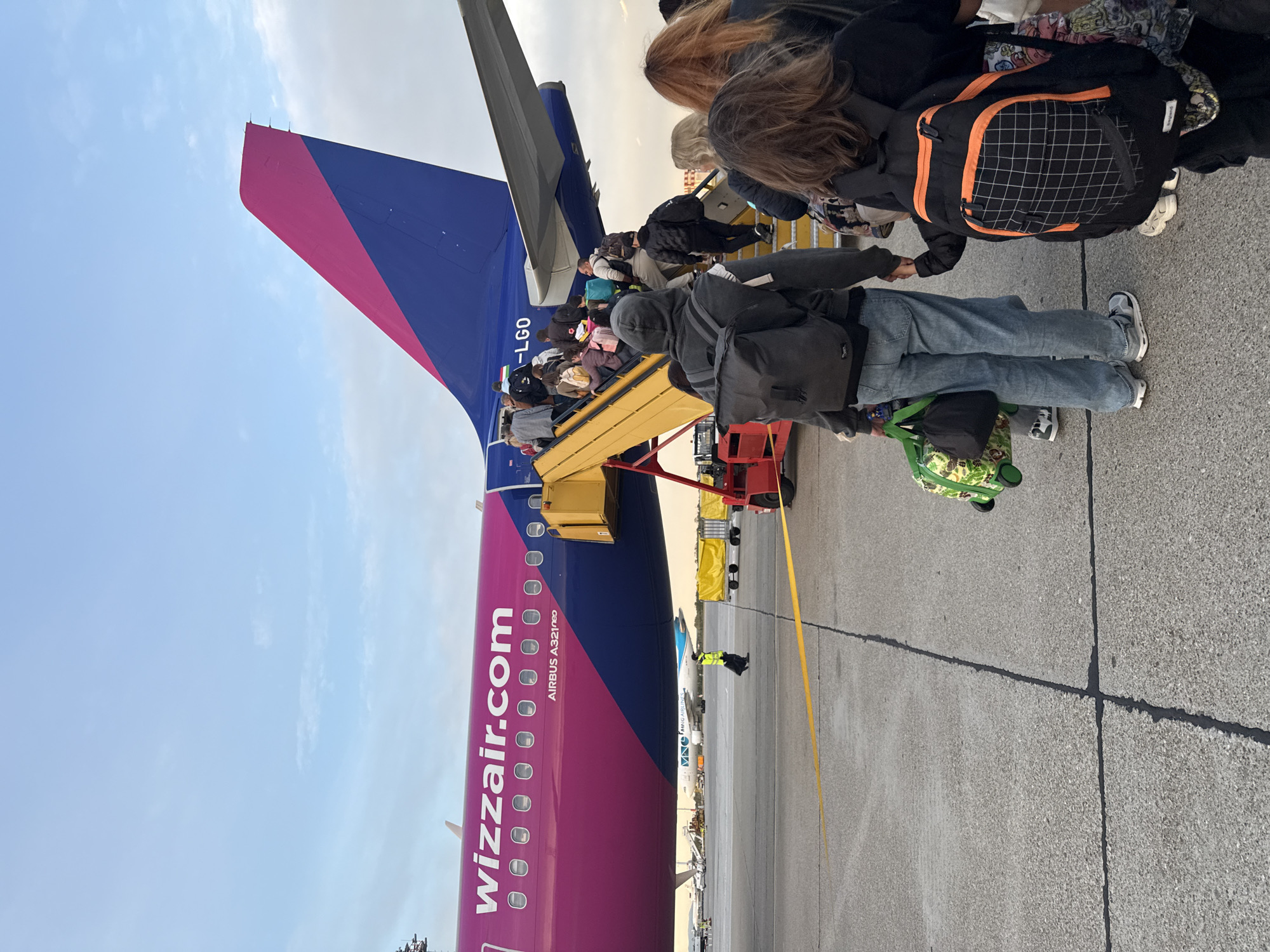 Passengers disembark a Wizzair Airbus A321neo, tail number HA-LGO, at Bratislava Airport in Slovakia. Passengers are exiting the aircraft via mobile stairs. A ground worker is visible near the plane's wing. The tarmac is concrete. The sky is overcast.