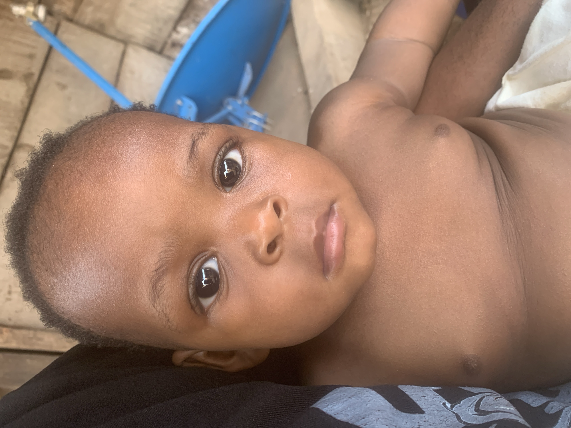 A close-up, high-angle shot captures a young baby, appearing to be an infant or toddler, lying on their back. The baby has a dark skin tone, dark, tightly curled hair, and large, deep brown eyes that gaze directly at the viewer with a curious expression. A bright reflection, possibly from a window or overhead light, is visible in each eye. The baby's lips are full and slightly parted. The upper body is bare, revealing smooth skin on the chest and arms. The baby appears to be resting on a dark-colored fabric, possibly clothing, which is visible in the lower left portion of the image.
In the background, out of focus, is a bright blue, dish-like object, resembling a small satellite dish or another piece of outdoor equipment. Below this object, blurred light-colored pavement or tiled ground can be seen. The lighting is bright and even, indicating daytime. The scene suggests an outdoor or semi-outdoor setting. No text is visible in the image. The image is oriented such that the baby's head is towards the left and their body extends to the right. The location context is Nkpor, Nigeria.