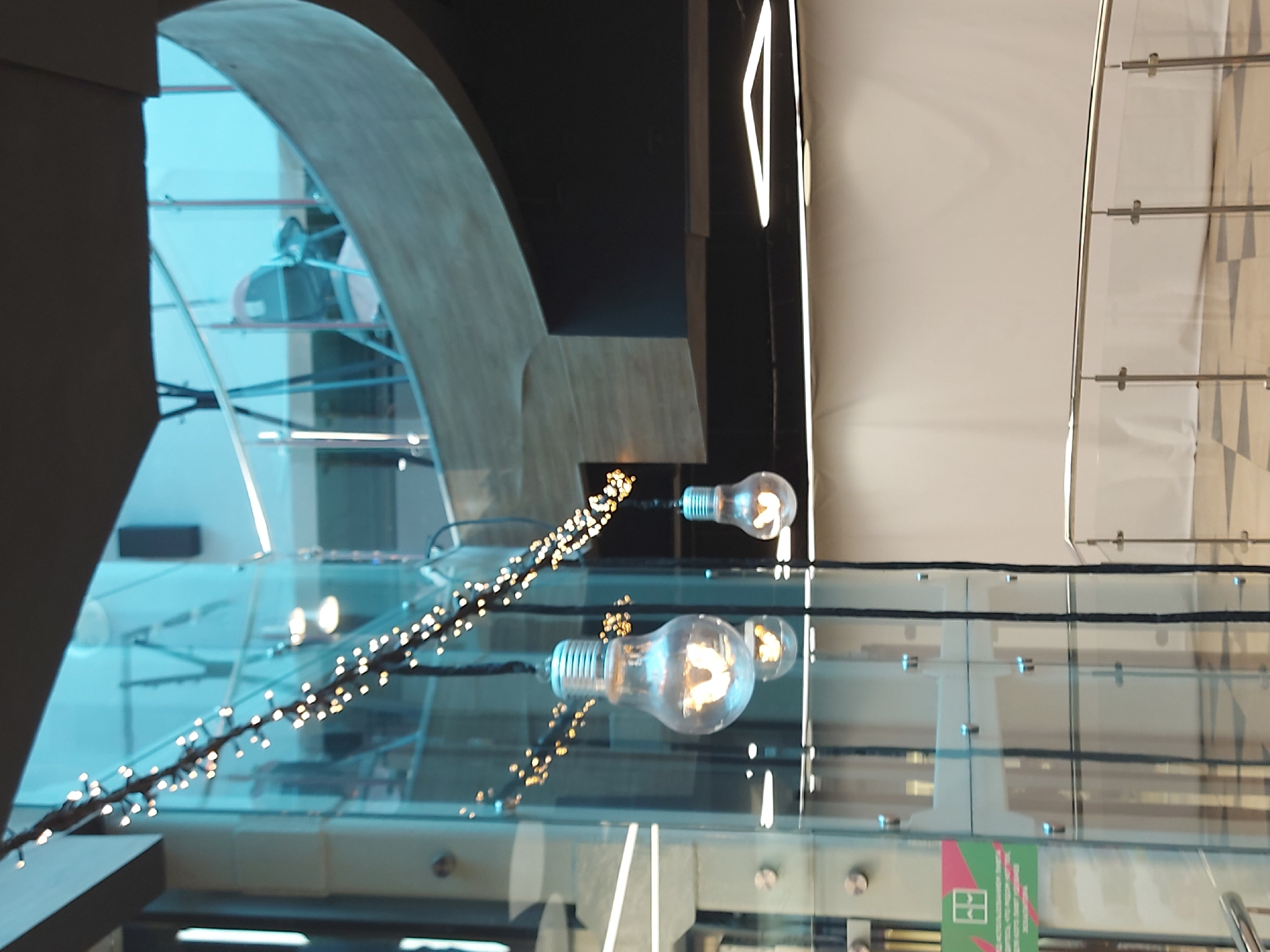 The image depicts an interior architectural scene in Saint Petersburg, Russia, featuring light fixtures and glass elements. The foreground showcases two exposed light bulbs suspended by a dark string of small lights. These appear to be reflected on a glass surface. The background includes architectural features like an archway and an escalator, suggesting a multi-level interior space, potentially a shopping mall or public building. One can spot people in the background, including one individual riding the escalator. The architectural design is modern, with elements of glass, steel, and neutral-colored walls.
