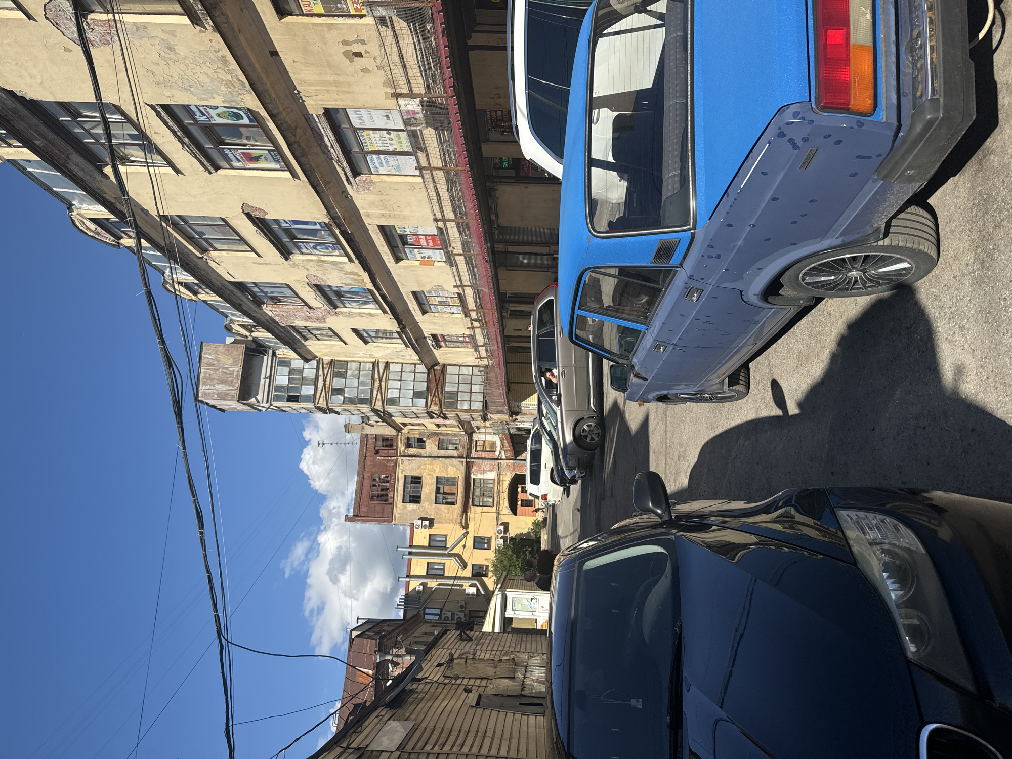 This is a medium-angle shot of a narrow street in Saint Petersburg, Russia, likely in an older, industrial district or courtyard. The buildings are aged, with peeling paint, exposed brick, and visible signs of wear. The architecture is varied, with some buildings featuring wooden siding and others with multi-story facades adorned with numerous windows. Wires are strung overhead, typical of urban infrastructure.
Several cars are parked or driving in the street, including a dark sedan in the foreground on the left, a blue sedan prominently in the center-right, a silver SUV further back on the right, and a white SUV behind it. There are also at least two pedestrians visible in the background near an entrance. One is a man in a light-colored shirt talking to another person. Another pedestrian, possibly a child, is walking further down the street. The sky is bright blue with scattered white clouds. The overall impression is of a functional, lived-in urban environment.