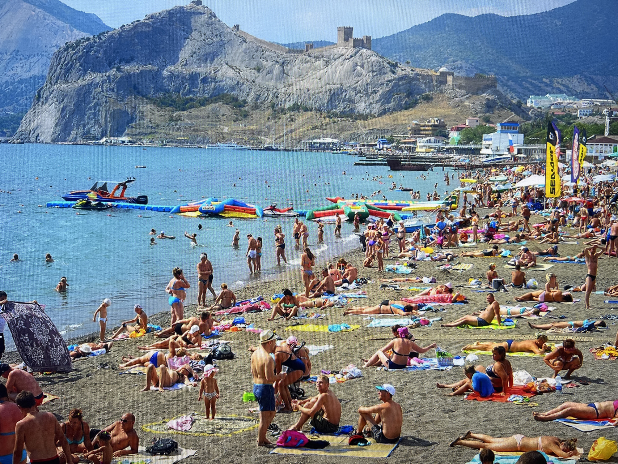 This is a wide-angle, eye-level shot of a crowded beach on a sunny day, with a mountain range and a fortified structure in the background. The beach is filled with people sunbathing, swimming, and engaging in water activities. Numerous colorful inflatable water toys are scattered in the shallow water near the shore. Jet skis are also visible in the water, suggesting recreational opportunities. In the distance, a town or city is built along the coastline, nestled at the base of the mountains. The sky is clear and blue. The scene depicts a bustling summer vacation spot.