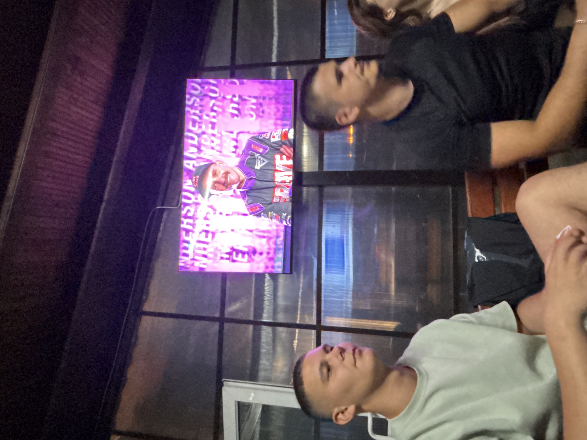 The image shows a group of people gathered at an outdoor venue, possibly a restaurant or bar, in Nedelino, Bulgaria. A large screen displays an image of a man in racing attire, identified by text on the screen as "ANDERSON". Two young men are seated in the foreground, facing the screen. The man on the left wears a light green t-shirt, while the man on the right wears a black t-shirt. The environment appears to be covered, with a slatted ceiling and translucent walls, suggesting an open-air structure. The lighting is dim, with the screen being the primary light source.