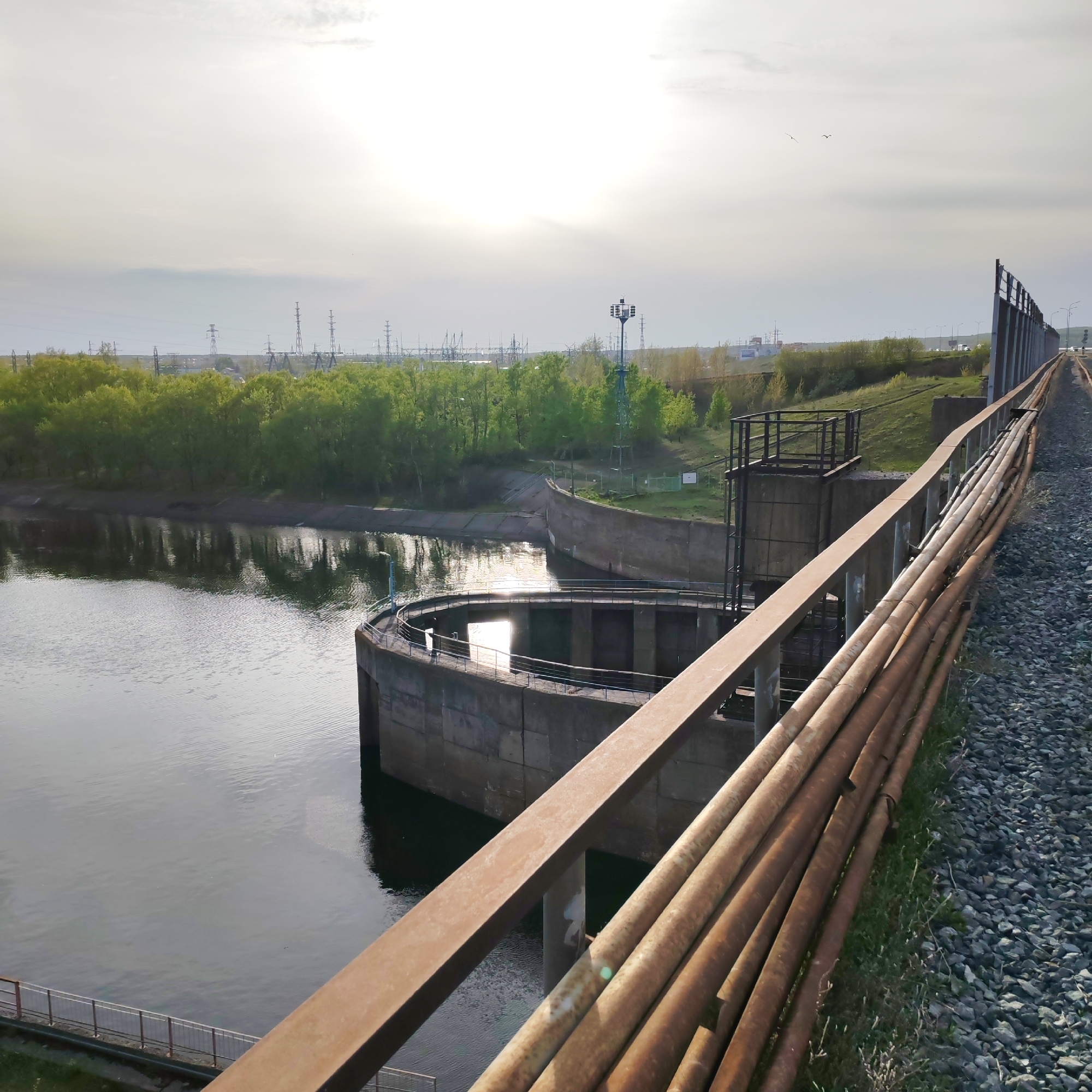 This image captures a view of the Kama River in the city of Chaikovsky, Russia. A concrete dam-like structure with a circular section is partially visible in the foreground. Above the dam and further along to the right, a rusty metal railing and a series of parallel pipes run alongside a track covered in grey gravel. Beyond the river, a line of trees, power lines, and a communications tower is visible under a cloudy sky with the sun shining through. There are no people depicted in the image.